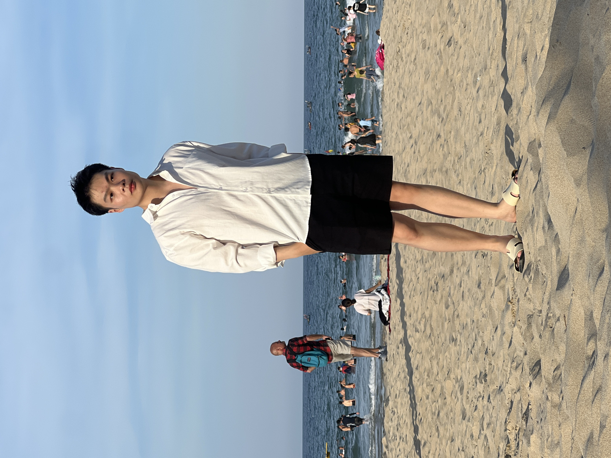 The image shows a beach scene in Da Nang, Vietnam. In the foreground, a young Asian man stands facing the camera, wearing a white unbuttoned collared shirt and black shorts. He has his hands in his pockets and is wearing light-colored sandals. The beach sand is a light beige color with visible texture. In the background, the ocean is a deep blue, with many people swimming and wading in the water. The sky is clear and a pale blue. To the left of the main subject, a man in a red and black plaid shirt and khaki shorts, carrying a teal backpack, is seen from behind, looking out at the water. Other beachgoers are scattered throughout the scene, some sitting on the sand and others in the water. The overall impression is a sunny day at a crowded beach.