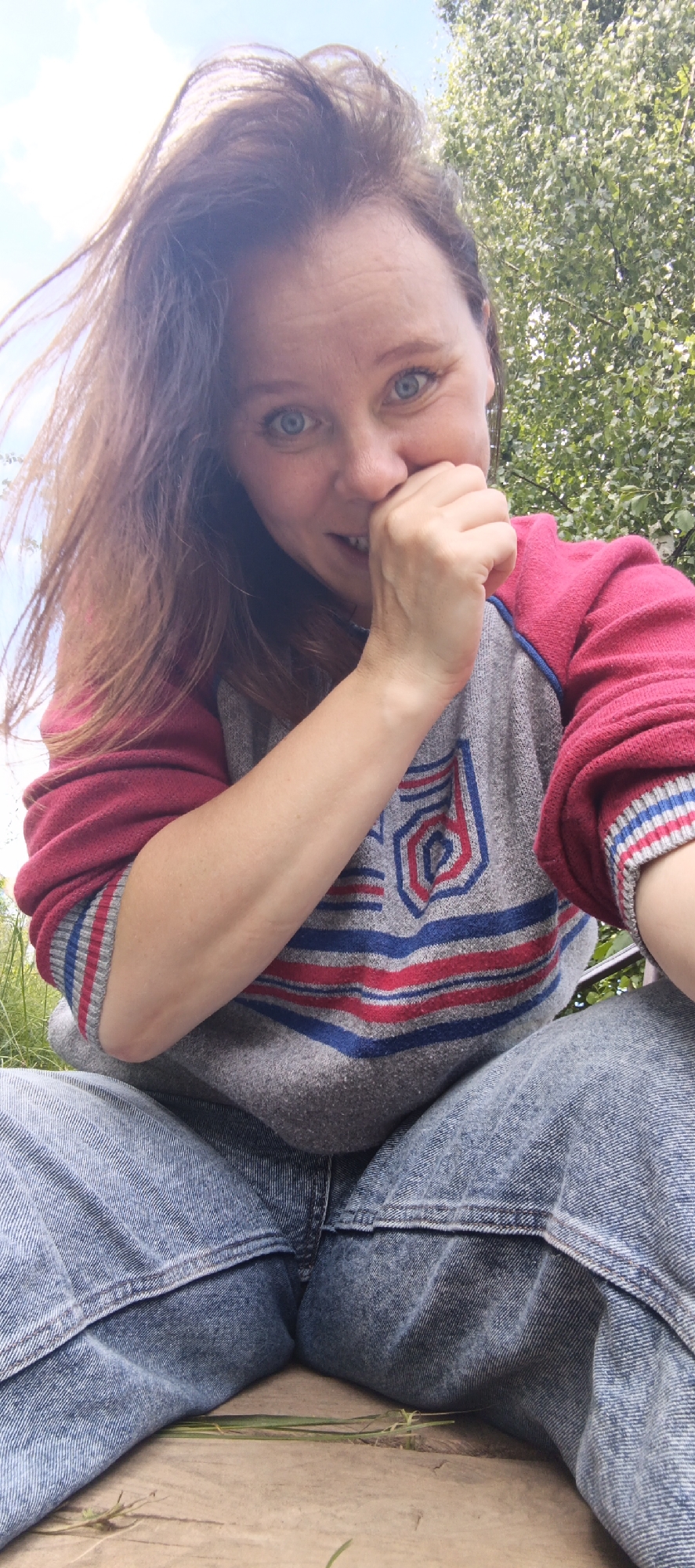 A low-angle, outdoor, medium shot captures a woman sitting on a wooden surface, possibly a bench, with grass and trees in the background, under a bright sky. She has long brown hair and blue eyes, and she is smiling and covering her mouth with her right hand. She is wearing a grey sweater with red and blue stripes and trim, and blue denim jeans. The woman appears to be in Chaikovsky, Russia, given the overall context.