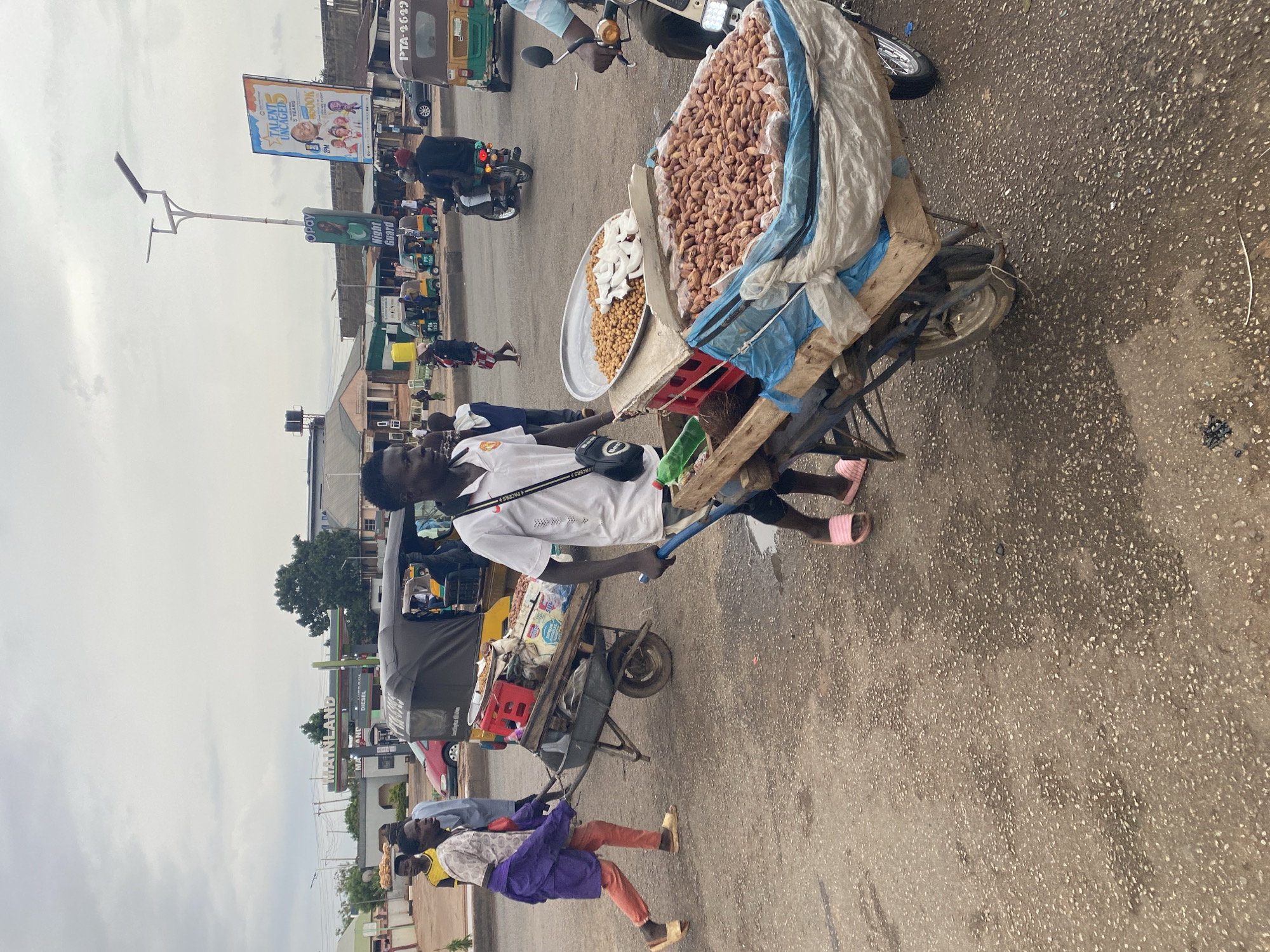 The image shows a street scene in Gura Top, Nigeria. A vendor pulls a heavily laden cart piled with peanuts and coconuts through a busy intersection. He wears a white shirt, dark shorts, and pink sandals. Behind him is a variety of vehicles, including auto rickshaws, and motorcycles. In the background are buildings with signs including "MAINLAND AND DIESEL" and an advertisement for "TALENT UNCAGED". Other people are seen walking nearby.