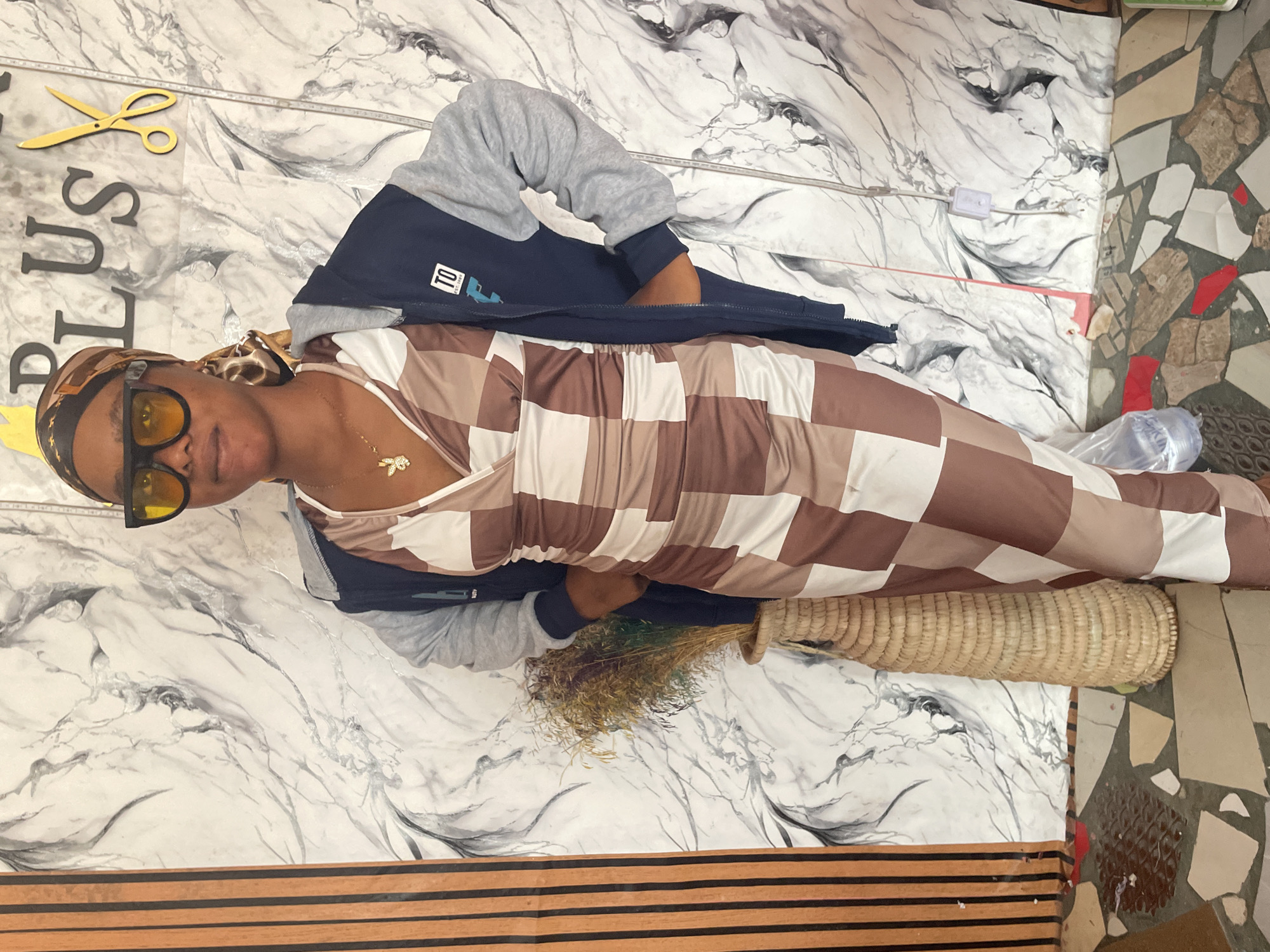 A woman is lying on a patterned surface, wearing a brown and white checkered dress, a grey and blue jacket, a gold headscarf, and yellow sunglasses. A woven basket and dried foliage are near her. Above her head, "PLUS" and a pair of scissors are visible on the surface. The background includes a mottled marble-like pattern and a mosaic tile floor, potentially indicating a location in Badagry, Nigeria.