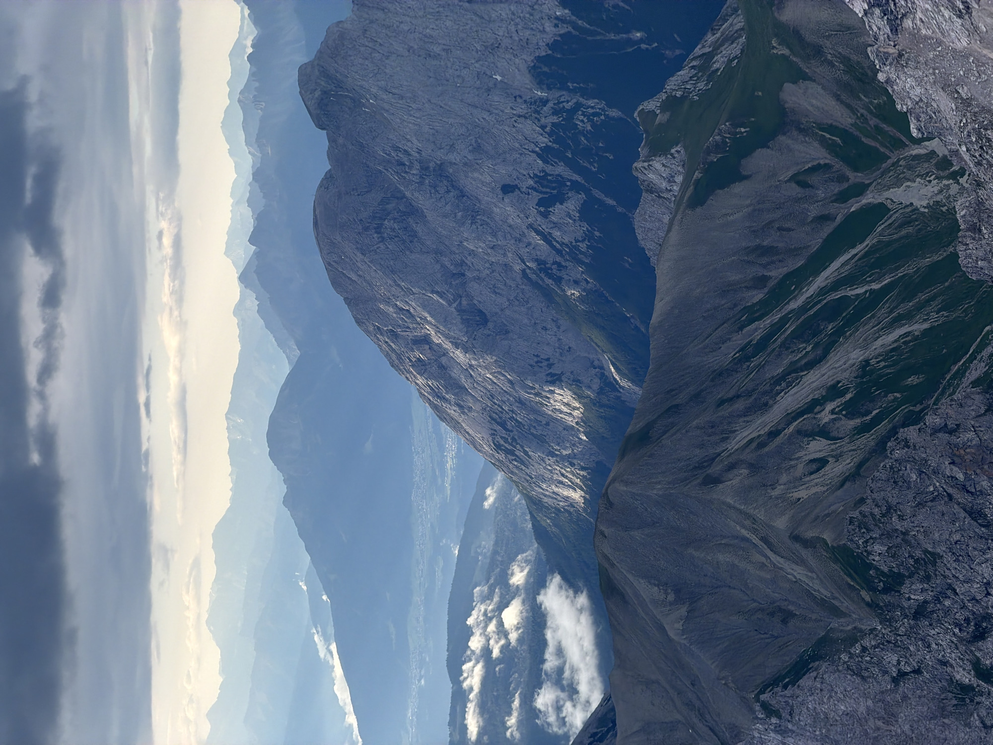 This is a high-angle, vertical shot of a vast mountain range, likely the Alps, with multiple layers of peaks receding into the distance. The foreground features rugged, gray, and dark green mountains with steep, rocky slopes. In the mid-ground, a prominent, large, light gray mountain with textured, weathered rock dominates the right side of the frame, catching the sunlight on its face. To its left, a valley is visible with some trees and scattered white clouds, and further in the distance, a hazy urban area or town with buildings is discernable. The background is a series of progressively fainter, bluish mountain ranges fading into the atmospheric haze. The sky above is filled with dramatic, dark gray clouds interspersed with patches of lighter sky, suggesting an overcast or transitioning weather condition. The scene captures a sense of immense scale and natural grandeur. The photograph is unlikely to be specifically located within the city of Garmisch-Partenkirchen itself, but rather depicts the surrounding mountainous landscape that characterizes the region. There are no people or animals visible in the image.