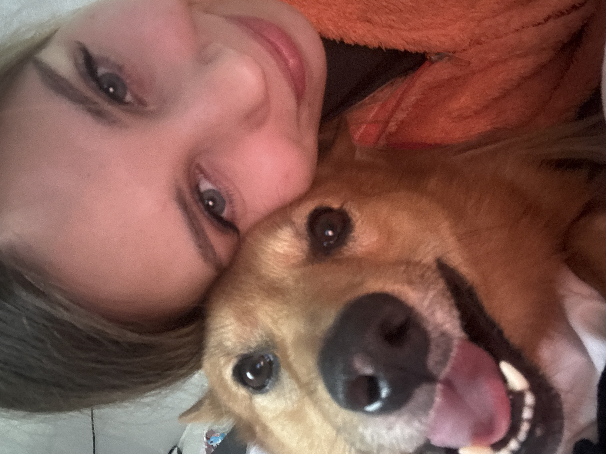 A close-up, low-angle selfie shows a young person and a dog. The person, likely female, is on the left side of the frame, with their head tilted towards the camera. They have fair skin, light brown hair, and blue eyes with visible eyeliner. Their lips are slightly parted and appear to be wearing lip gloss. They are wearing an orange, fuzzy jacket.
On the right side of the frame is a medium-sized dog with golden-brown fur. The dog's head is resting close to the person's head, and its mouth is open, with its pink tongue visible. The dog's eyes are dark and appear to be looking at the camera. The overall expression of both the person and the dog suggests affection and happiness.
The background is mostly out of focus, but hints of a white surface, possibly a blanket or bed, are visible. The lighting suggests an indoor setting, possibly during the day. The close proximity and shared space between the person and the dog indicate a close bond. No text is visible in the image. The scene is intimate and personal, capturing a moment of connection between a person and their pet.