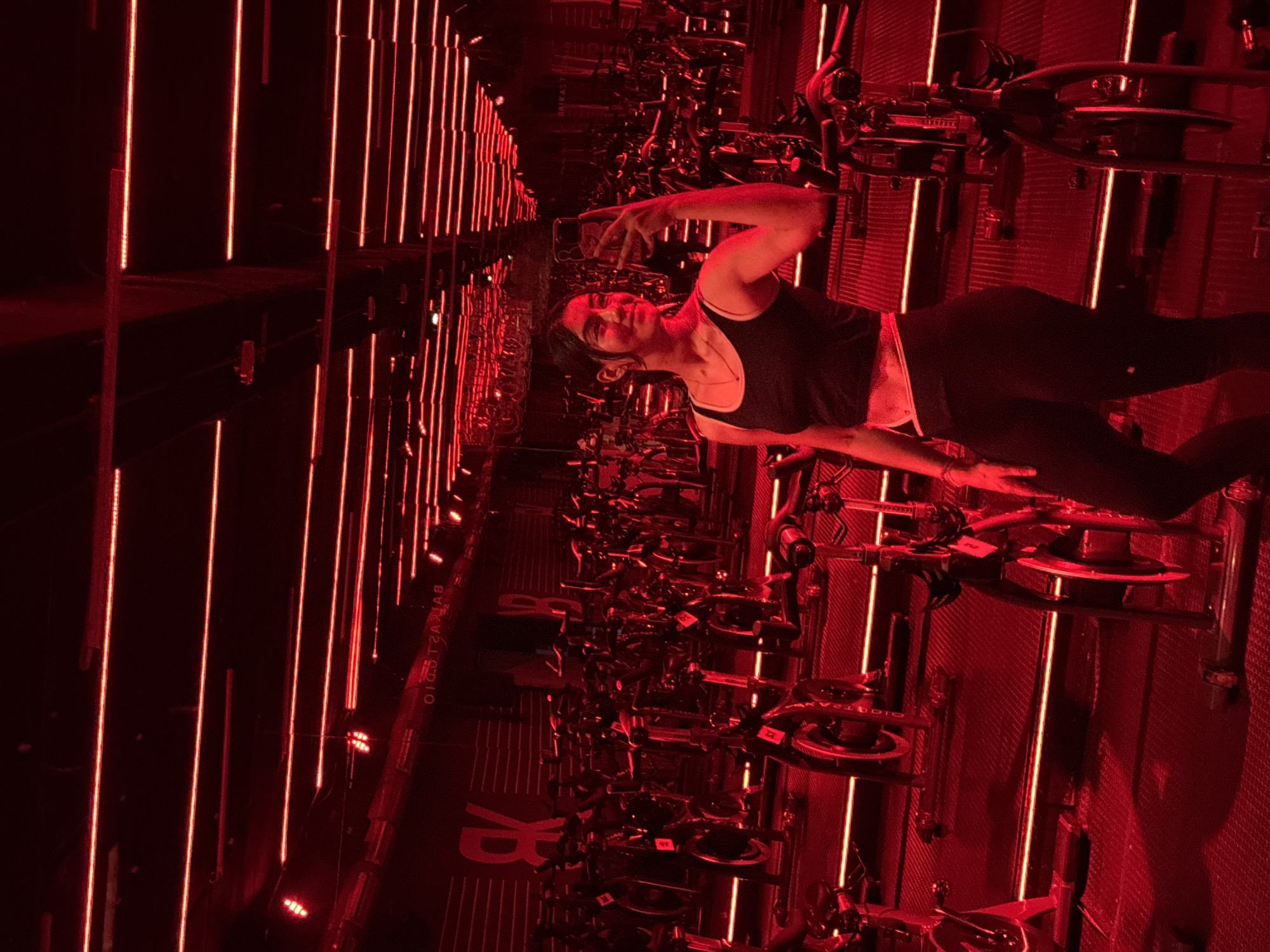 A woman poses for a photo in a dimly lit indoor cycling studio. The studio, likely located in Puebla, Mexico, is illuminated with red vertical lights. She wears a black tank top, dark leggings, and a necklace. The studio contains rows of stationary bikes with the word "RHYTHM" visible on the floor. The woman is striking a pose with one arm raised.