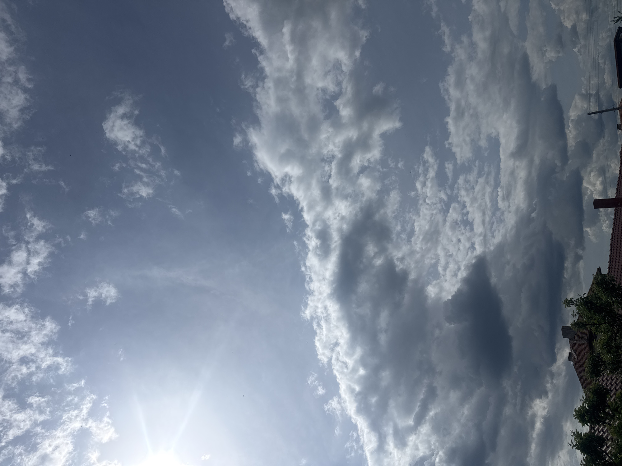 The image shows a sky with clouds. The sun is visible in the lower left corner. To the right of the image, there are portions of buildings and foliage. The location is Sarnevo, Bulgaria.