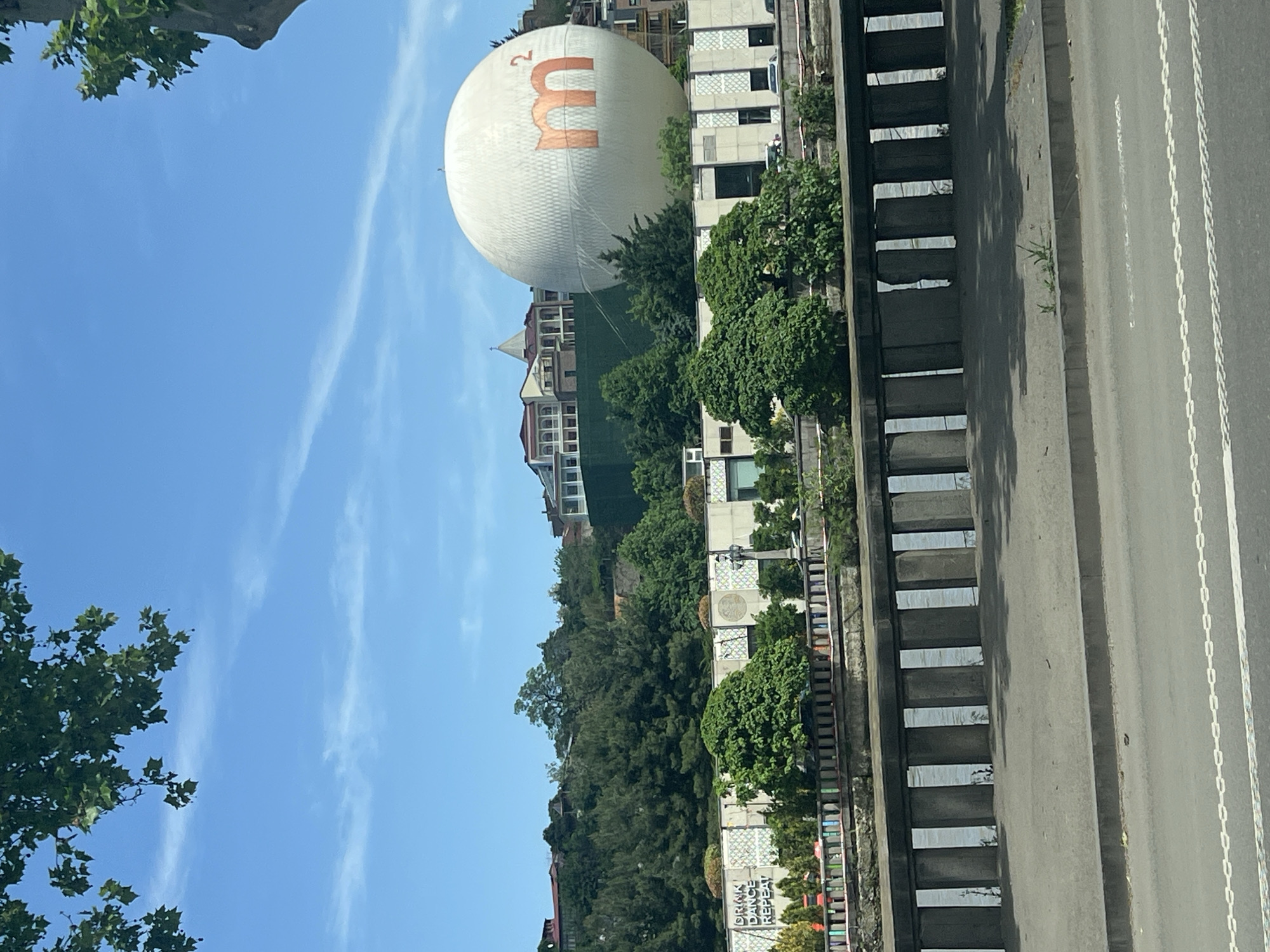 The image shows the city of Tbilisi, Georgia, with a prominent white balloon bearing an orange "M" logo hovering above buildings and trees. The sky is blue with light, wispy clouds. A multi-story building with visible windows is located to the right of the balloon. Lush green foliage is present in the midground. A roadside barrier and a section of gray asphalt road with white lane markings run along the bottom right of the frame. The view is from an elevated position looking downwards. "DRINK DANCE REPEAT" is written on a concrete wall. A building with a red roof is visible in the background, below the balloon.