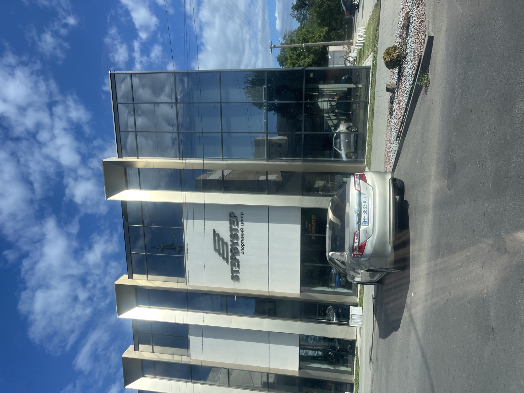 The image shows the front facade of the "Step By Step" sports center in Uzhhorod, Ukraine, under a bright blue sky with scattered white clouds. The building features a modern design with large glass windows and a prominent white and beige exterior. The "STEPBYSTEP" logo and the Ukrainian text "спортивний центр" (sports center) are clearly visible above the entrance. A silver sedan is parked in front of the building on an asphalt road, with its rear license plate readable as HH 3835 AH. A landscaped area with gravel and a small bush separates the road from the building's entrance. The scene appears to be a typical daytime view of a commercial establishment.
