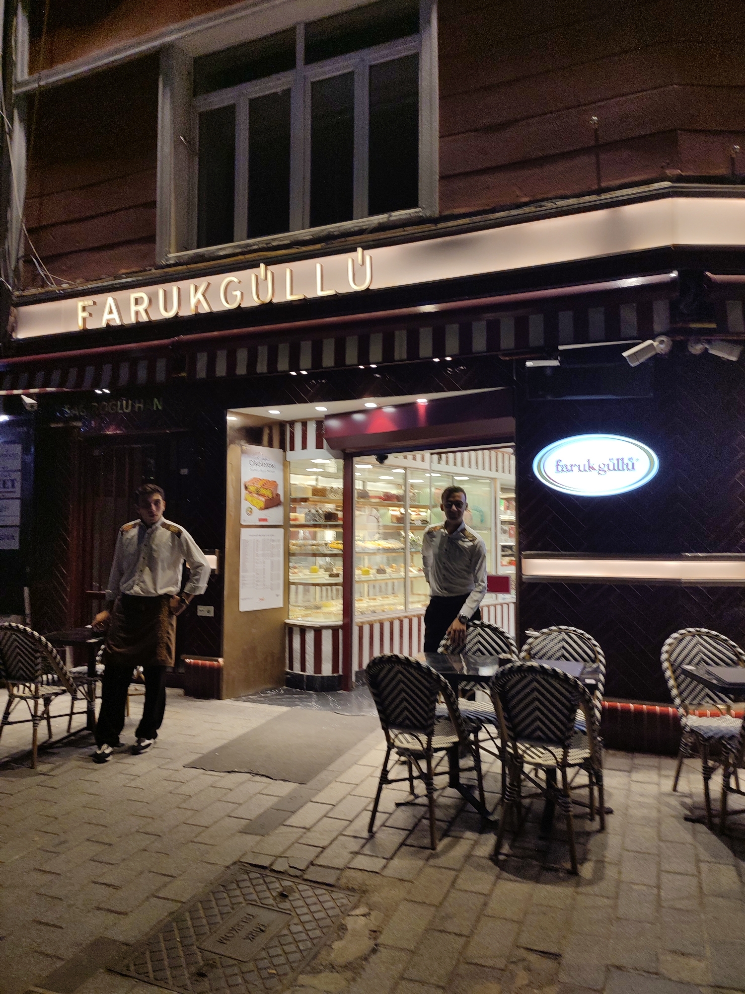 In Beyoğlu, Turkey, a brightly lit patisserie named Faruk Güllü is captured at night. The exterior features a prominent illuminated sign with the name "FARUKGÜLLÜ" in large, white, sans-serif letters. Below the sign, a striped awning in dark red and white marks the entrance to the shop, where a display of pastries is visible through a glass storefront. Two male employees, dressed in white shirts and dark trousers, stand near the entrance. One employee is positioned inside the patisserie, partially obscured by the display cases, while the other stands outside, to the left of the entrance, with his hands on his hips. A few tables with wicker chairs are arranged on the cobblestone sidewalk in front of the shop, illuminated by the ambient light. Security cameras are visible mounted on the building.