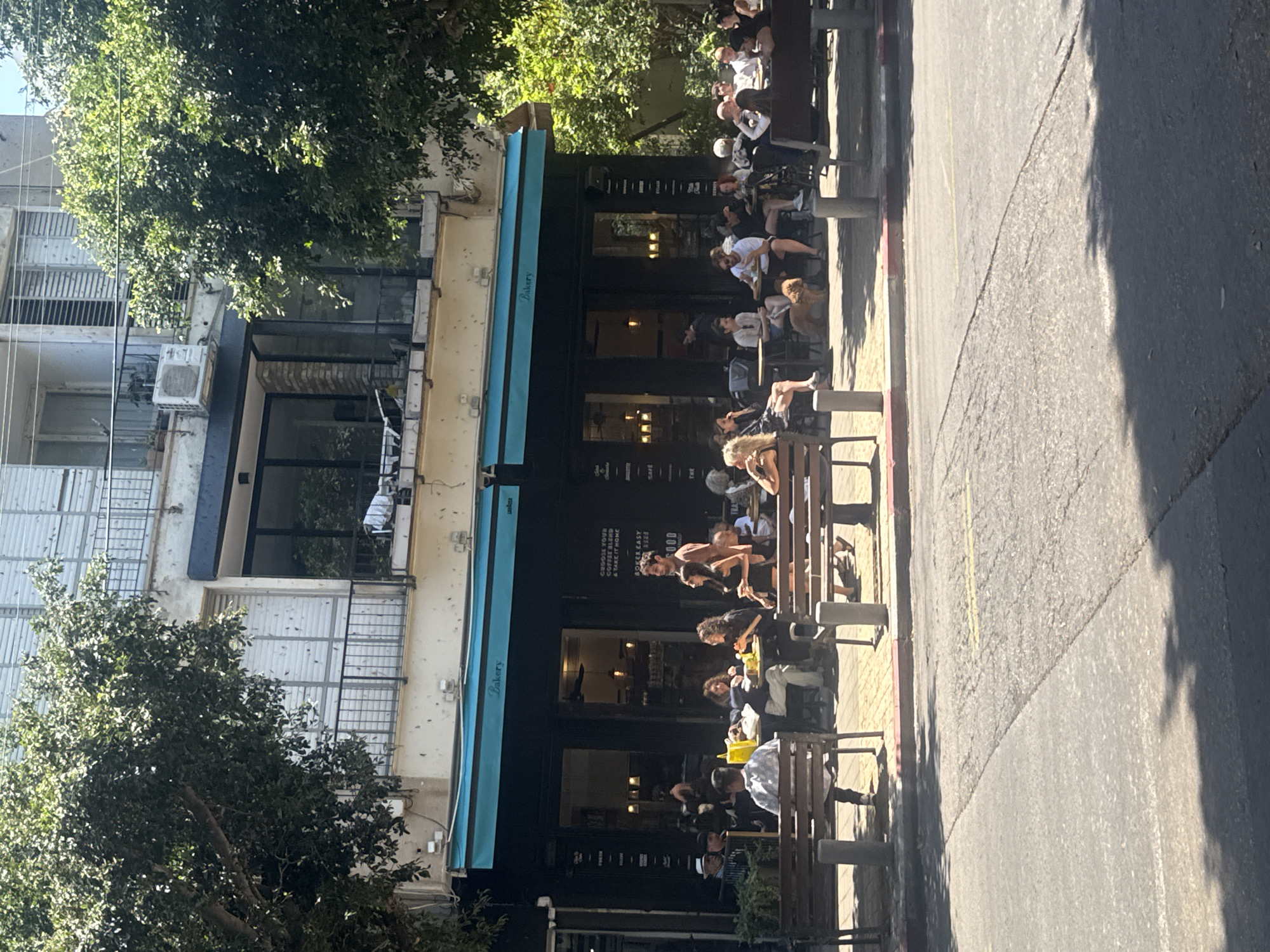 The image depicts the exterior of a bakery located in Tel Aviv, Israel. The bakery has a dark facade with a blue awning labeled "Bakery." People are seated at tables and benches along the sidewalk outside the bakery. The building next to the bakery is multi-storied with window bars and an air conditioning unit. Trees are visible on both sides of the buildings. The street is asphalt and shows a shadow from a nearby tree. The scene shows a bright, sunny day.