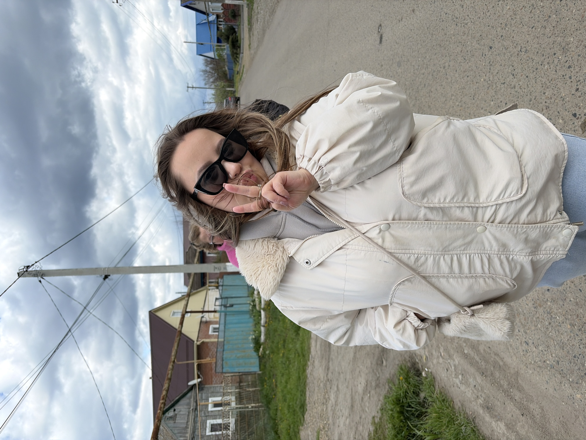A woman with light brown hair and dark sunglasses is standing on a street in an unidentified city. She is wearing a light-colored jacket with a fur-lined collar and is holding up two fingers in a peace sign. In the background, there are houses, utility poles, and an overcast sky. The ground is paved, and there is some grass growing along the side of the road.