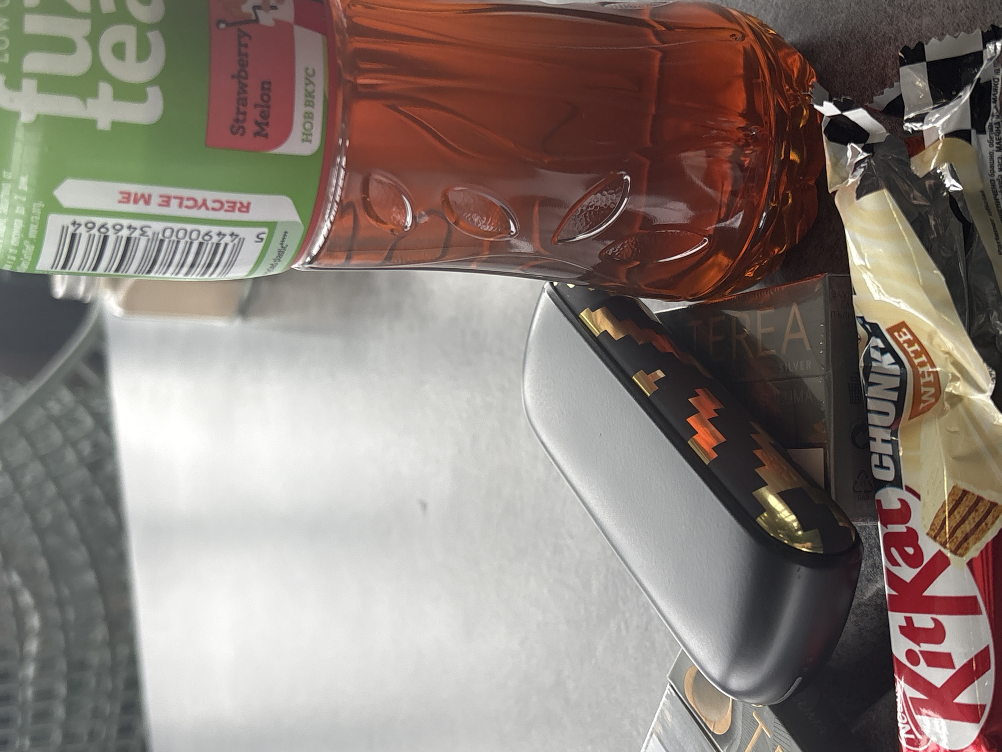 A close-up, still-life photograph shows a bottle of Fuze Tea with a strawberry-melon flavor lying on its side. Next to it is a pack of Terea Silver cigarettes, a Kit Kat White Chunk candy bar, and a black device with gold detailing. The photograph appears to have been taken indoors on a grey surface. The presence of Bulgarian text ("НОВ ВКУС", "пълн", "БРАШНО") indicates that the location might be in Bulgaria, possibly Sofia.