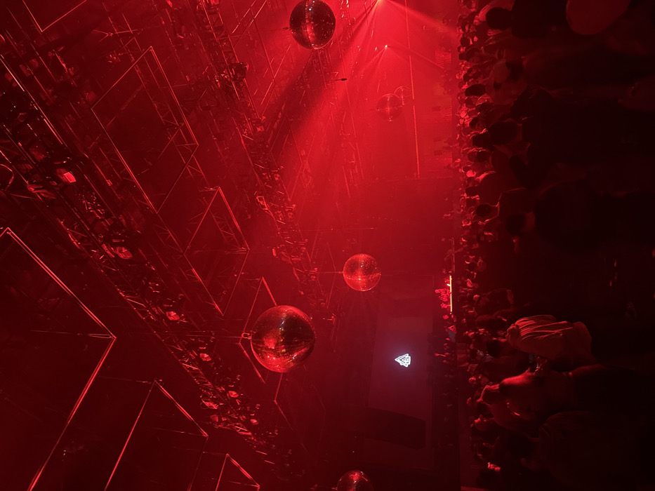 An intense red light floods the scene, illuminating hanging disco balls in varying sizes. The ceiling is structured with mirrored geometric patterns. To the right, a wall appears textured with organic material. A soft glow emanates from a logo near the wall, adding a focal point amidst the vibrant red ambiance.