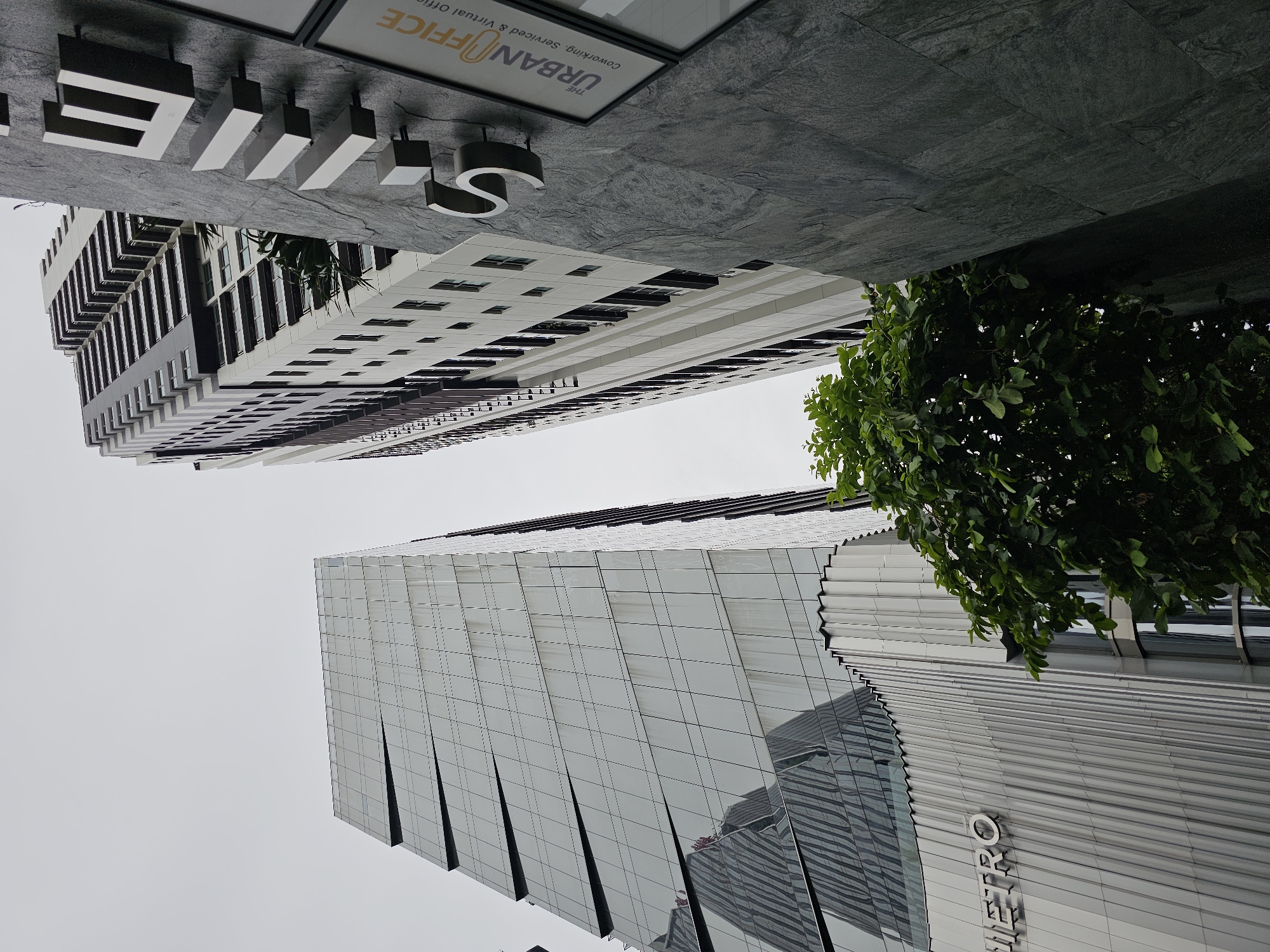 This is a low-angle shot of modern buildings in Bangkok, Thailand. In the foreground, a building with a rippled metallic facade has the word "METRO" in raised silver lettering. To the right of this building, a taller skyscraper with a grey stone exterior displays large, metallic letters spelling "S" and partially visible letters from "THE URBAN OFFICE" on a sign. Behind these structures, a very tall, slender building with rows of balconies stretches towards the sky. A lush green tree with broad leaves is positioned in the lower right quadrant of the image, partially obscuring the view of the buildings. The sky is overcast and a pale grey.
