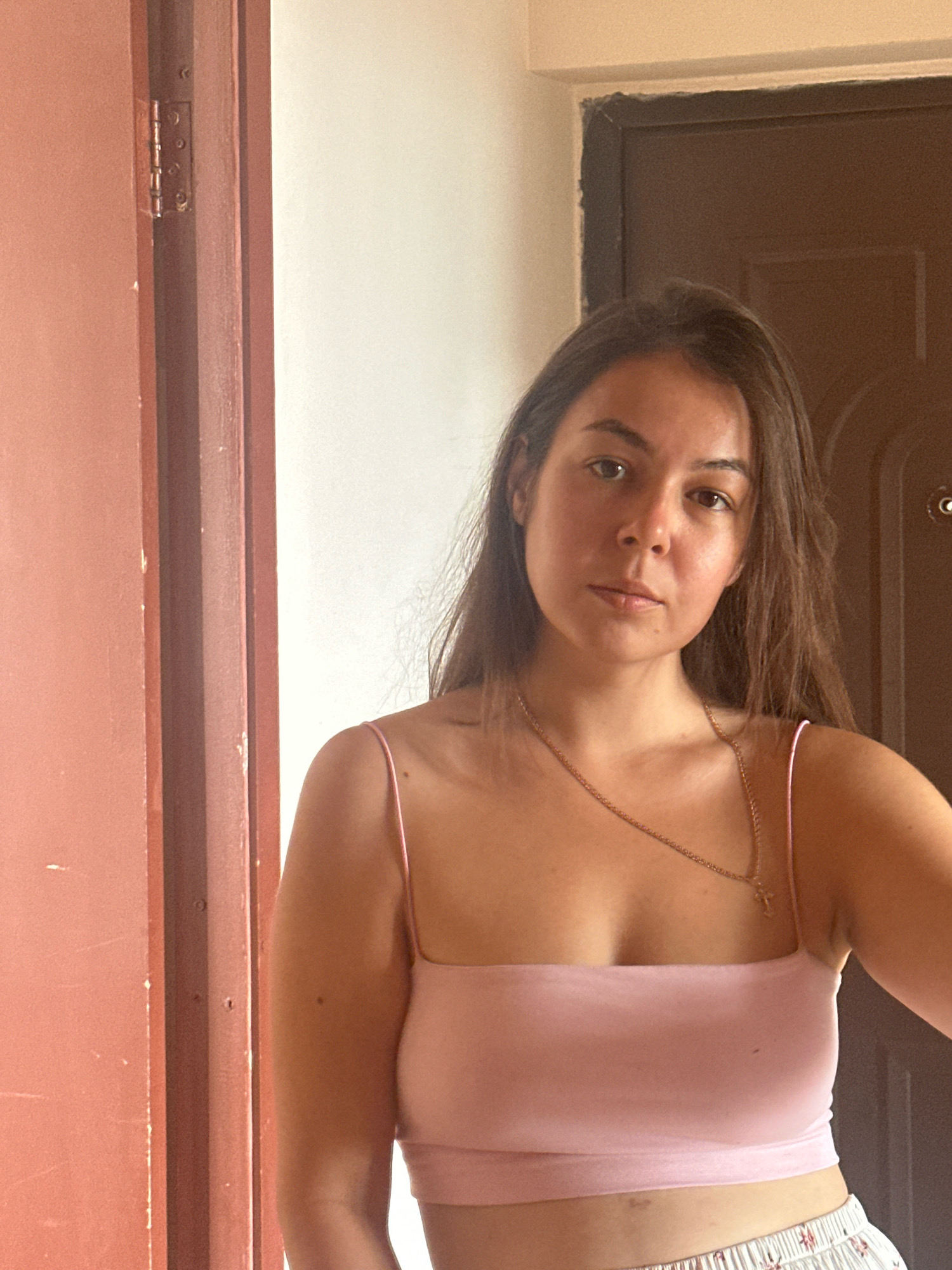 A woman with long brown hair stands in front of a door frame, in what appears to be an interior space in the city of Gyumri, Armenia. The woman is wearing a pink, spaghetti strap crop top and patterned shorts. She has a gold necklace with a cross pendant. The door frame is painted a dark brown and appears to be made of wood. To the left of the woman is a reddish-brown door with visible hinges. The background wall is a pale yellow. The lighting suggests it is daytime, with light coming from an unseen source to the right.