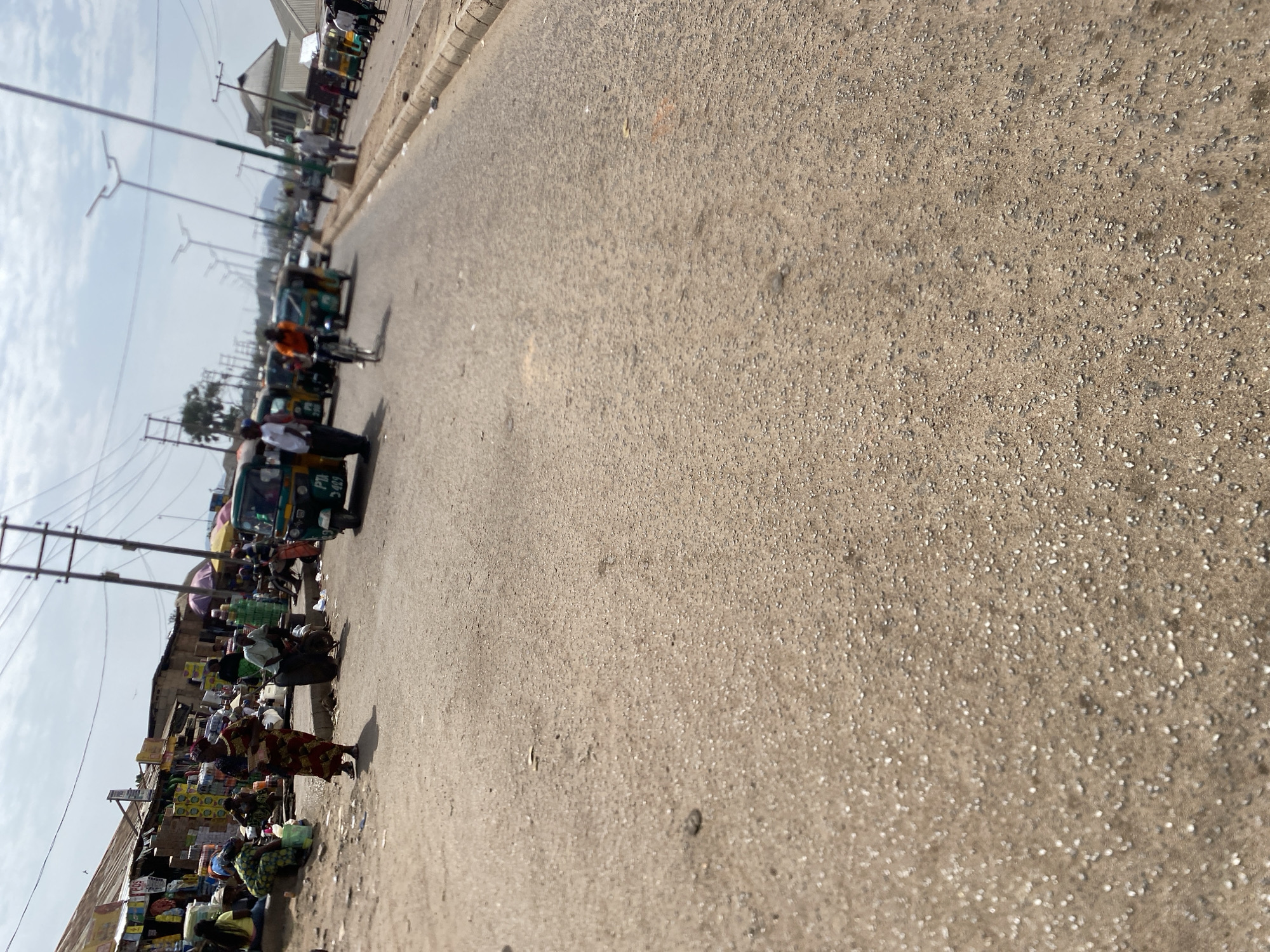 The image depicts a street scene in Bukuru, Nigeria. The road, made of a coarse asphalt or concrete, takes up most of the frame and recedes into the distance. On the left side of the road, there is a line of people, likely vendors, sitting or standing next to their wares. The merchandise appears diverse, possibly food items and other goods. Several "keke" or auto-rickshaws are parked further down the street. Overhead power lines are visible. The sky is partially cloudy.