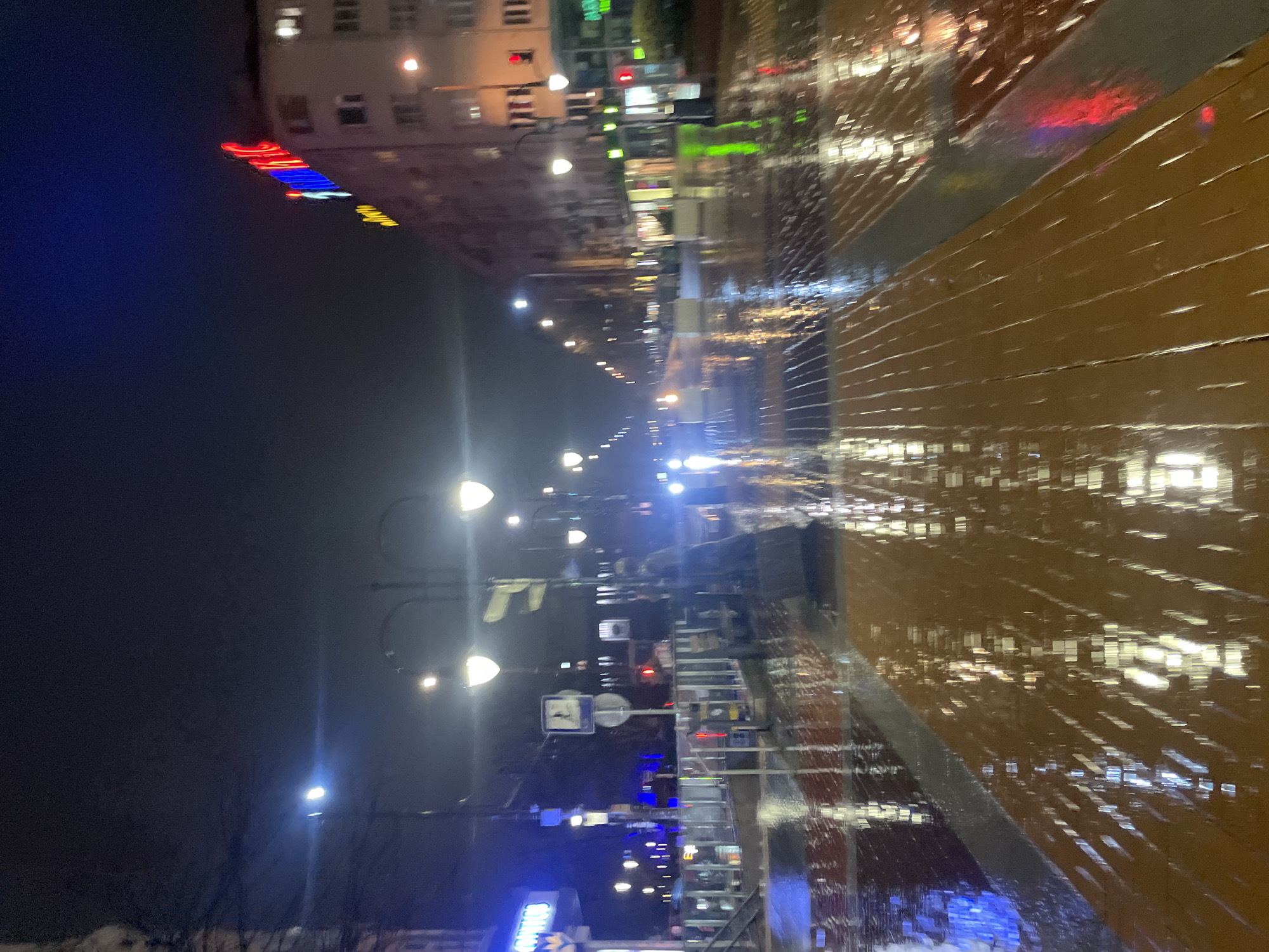 The image depicts a nighttime street scene in Sofia, Bulgaria. The street is wet from rain, and the reflections of lights are visible on the pavement and surrounding surfaces. There are buildings on either side of the street, illuminated by streetlights and neon signs. A row of streetlights extends into the distance, with their light scattering through the rainy atmosphere. The street appears to be empty of people and vehicles.