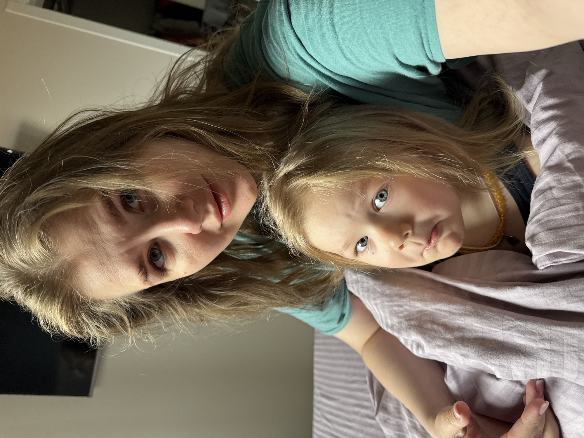 Two individuals, an adult and a child, are close together. The adult is wearing a green top. The child appears to be wearing a necklace. They are positioned on a bed covered with lavender-colored bedding. The background includes a light-colored wall and part of a dark object, likely a television or a piece of furniture. No location is visible. The image has been rotated.