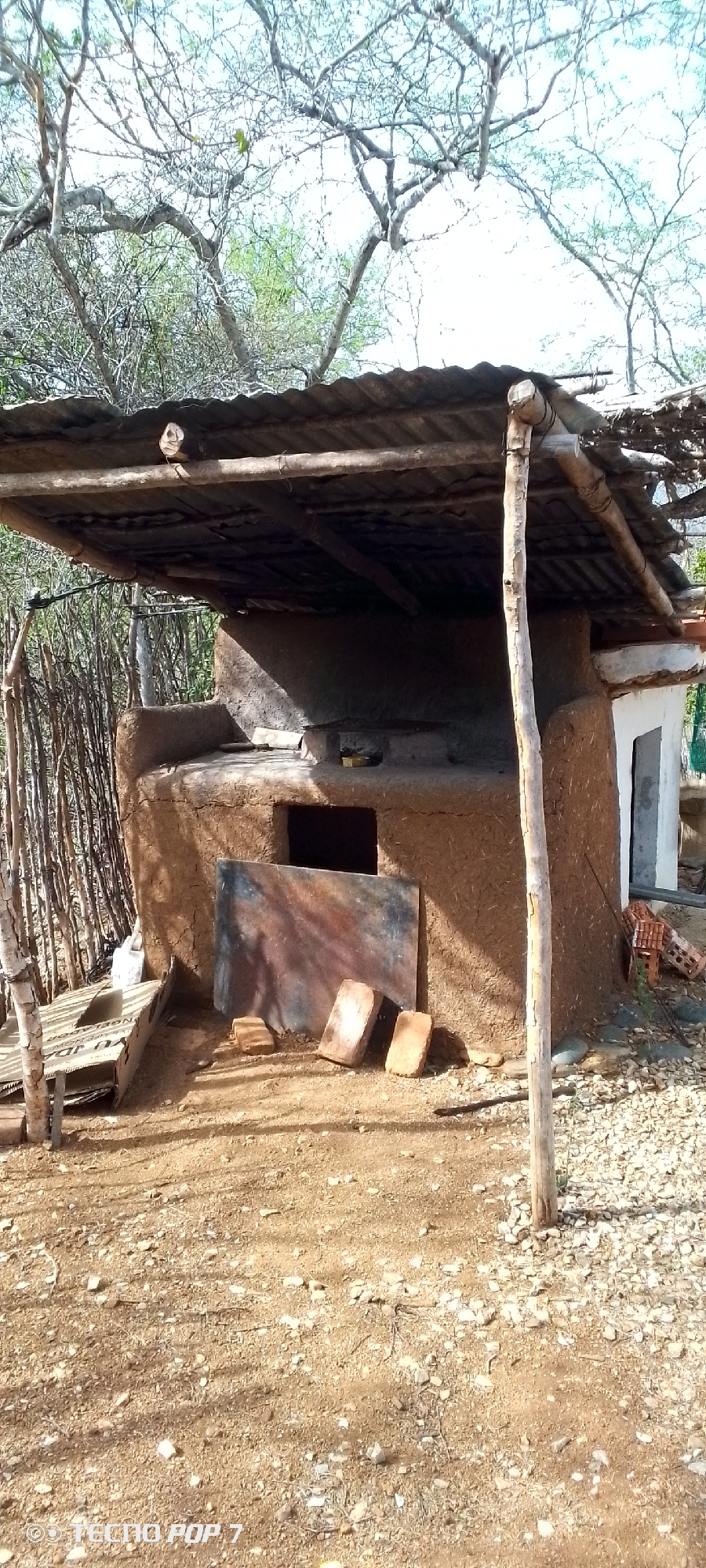 An outdoor scene depicts a rustic, mud-built structure, likely a wood-fired oven or kiln, situated in a dry, unpaved area. The structure is covered with a corrugated metal roof supported by wooden beams and poles. In front of the oven, a large, weathered piece of sheet metal rests on the ground, flanked by two bricks. To the left of the oven, a low wooden bench or platform is visible. The surrounding environment includes sparse vegetation, a fence made of branches, and a clear sky overhead. The ground is covered with dirt, small stones, and shadows cast by the trees. The image is marked with the text "© TECNO POP 7".