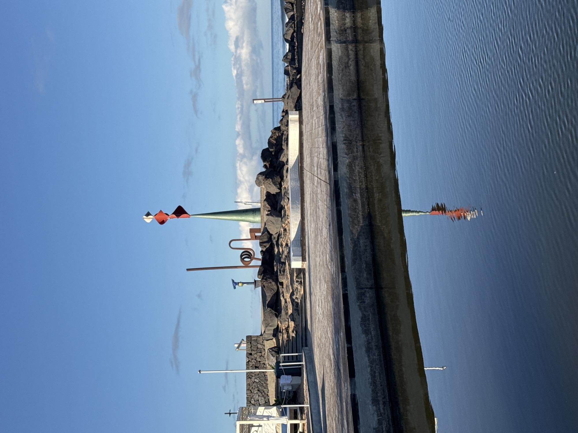 The image depicts an outdoor scene in Arona, Spain, likely a waterfront promenade or park. In the foreground, a calm body of water reflects the sky and a portion of the scene. The water surface exhibits small ripples. Beyond the water, a concrete walkway leads to an elevated area with dark volcanic rocks. A modern sculpture featuring a green conical base topped with red and white diamond-shaped elements is prominently visible on the right side of the elevated area. Various other sculptural elements, including a brown spiral and a chair-like structure, are also present. A white beach cabana with loungers is situated on the left side of the image, near some steps leading up to the rocky area. A white pole, possibly a flagpole or lighting fixture, stands near the cabana, accompanied by a small green trash receptacle and a white rope barrier. The sky is blue with scattered white clouds, and the ocean is visible in the background with gentle waves. The scene appears to be bathed in natural sunlight. There are no people visible in the image.