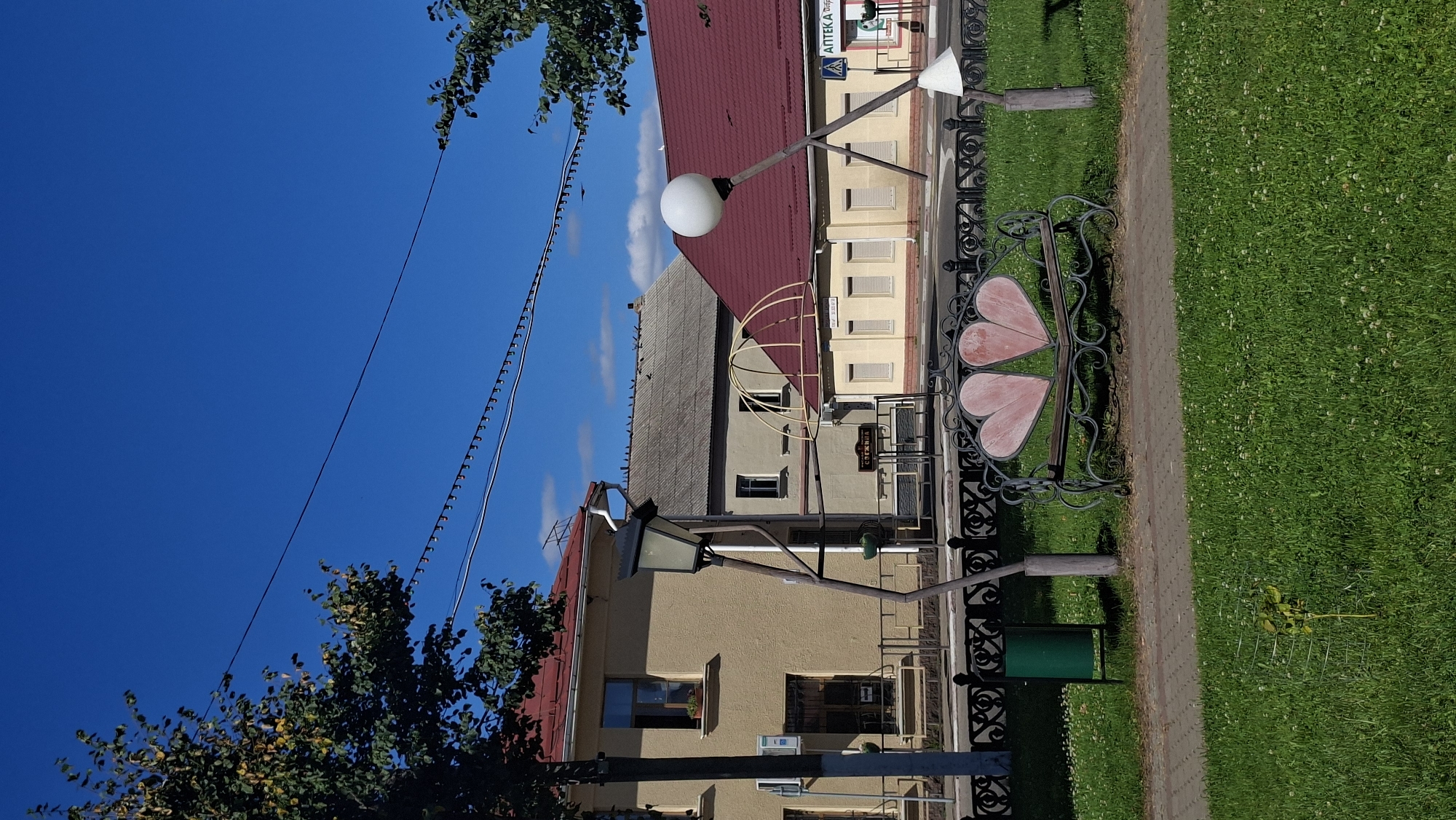 A medium shot captures a street scene in Pastavy, Belarus, under a clear blue sky. In the foreground, a park bench shaped like two hearts sits on a grassy area. To the left of the bench is a green trash can. Beyond the bench, a decorative black metal fence separates the grassy area from a paved walkway. Further back, two street lamps with unusually shaped poles stand on either side of the walkway. One lamp has a square-shaped fixture, while the other has a round, spherical light.
Behind the lamps and fence, a series of buildings line the street. The closest building on the left is a two-story structure with a light yellow facade and a red roof. To its right, another building with a gray roof has numerous birds perched on its peak. Further down the street, more buildings with red and yellow facades are visible, with one having a sign that appears to say "АПТЕКА" (Pharmacy). Overhead, strings of lights are strung across the scene, and trees with green foliage are visible on the left and right sides of the frame. The overall impression is a quiet, sunny day in a small town.