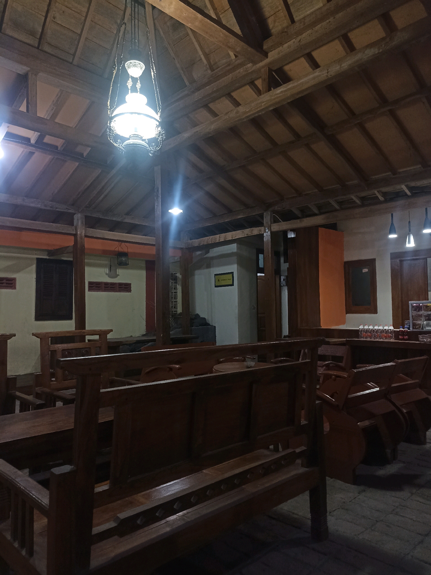 The image depicts the interior of a traditional wooden building, likely a restaurant or guesthouse, in Bakalanpule, Indonesia. The architecture features exposed wooden beams and rafters forming a pitched roof. In the foreground, there are several wooden benches and tables, suggesting a seating area. The lighting is dim, with a prominent antique-style hanging lamp illuminating the center of the space. Other light sources appear to be modern ceiling lights. The walls are painted in light colors, with some darker wooden accents and windows. In the background, there are additional seating areas, some with cushioned chairs. A counter area with bottles and what appear to be food items is visible on the right. The overall impression is one of rustic charm and a warm, inviting atmosphere.