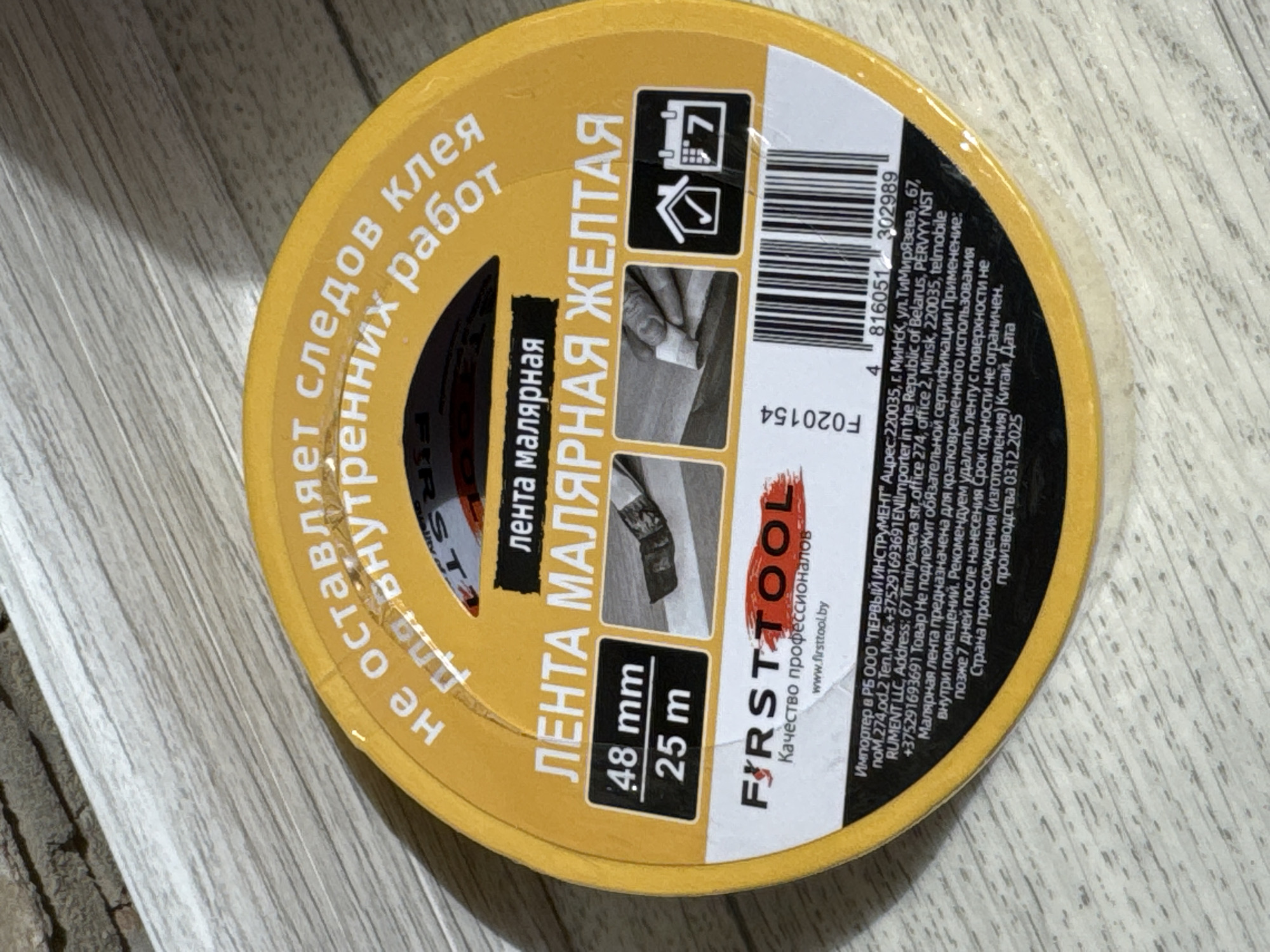 The image is a close-up, top-down shot of a roll of yellow masking tape. The tape is round and yellow with text and graphics printed on it. The text on the tape is in Russian and indicates it is "MASKING TAPE YELLOW". It also shows the dimensions "48 mm" and "25 m". The brand name "FIRST TOOL" is prominently displayed. There are also icons of a house with a checkmark and a calendar with the number 7. A barcode is visible at the bottom of the roll. The tape is lying on a light-colored, wood-grain surface. The context of "Яромінскі сельскі Савет, Belarus" is not directly visible in the image but refers to the location of the importer mentioned in the text on the tape, "PERVVY NST RUMENT LLC".
