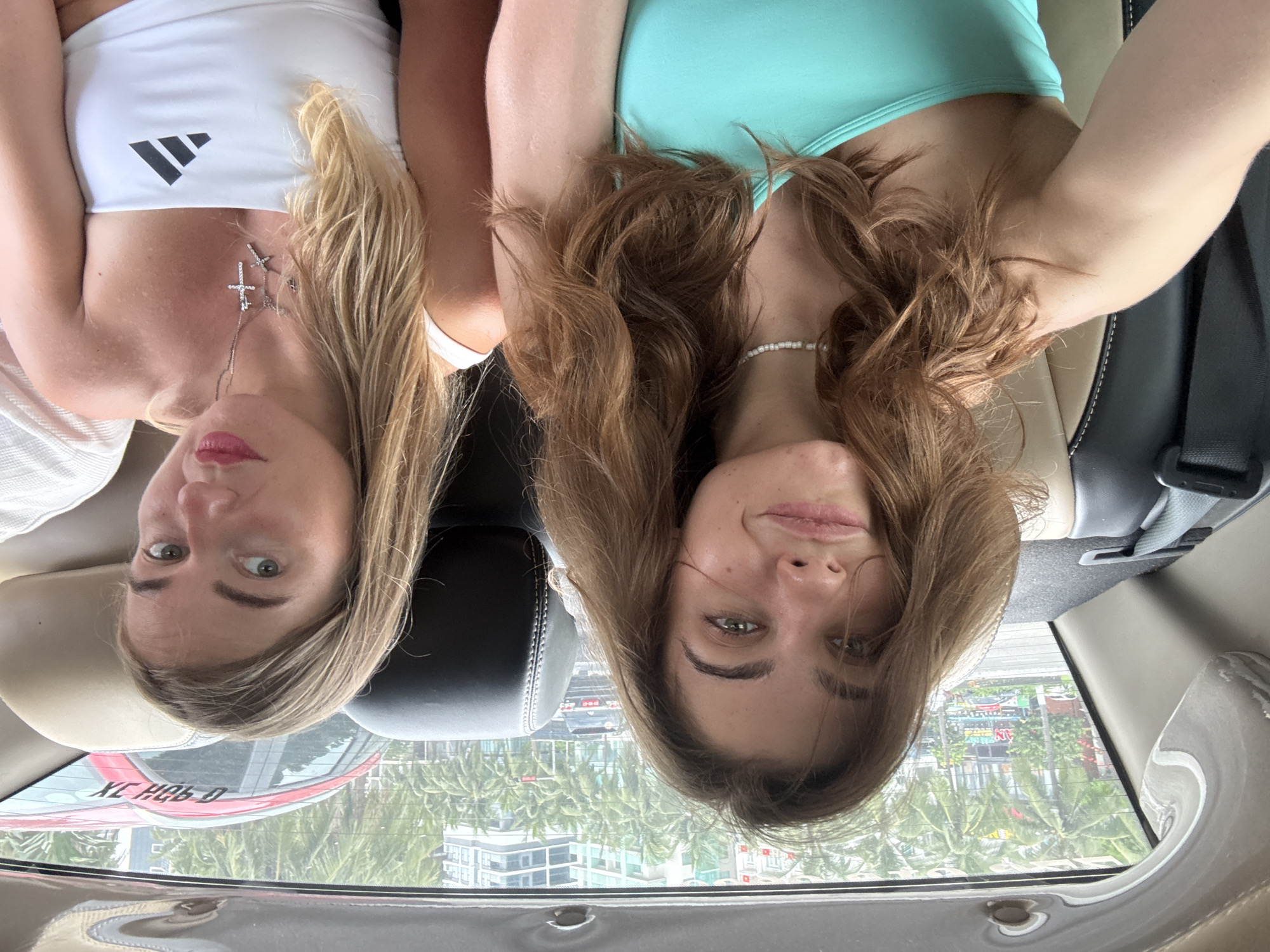 Two women are sitting in the backseat of a car. The woman on the left is wearing a white Adidas tank top and a silver necklace with a cross. The woman on the right is wearing a teal tank top and a pearl necklace. The car appears to be in Đà Nẵng, Vietnam, based on the view through the car window featuring palm trees, buildings, and a street with signage. The image is oriented upside down. A red car is visible through the rear window, with a partially visible license plate.