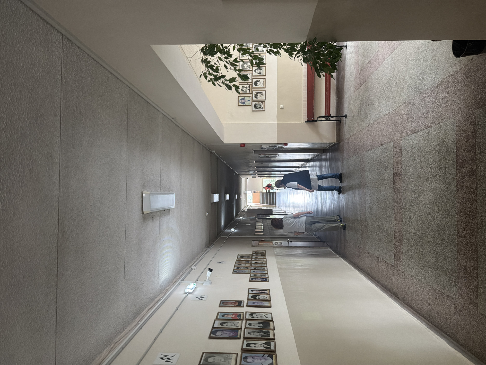 This image captures a long, well-lit hallway extending into the distance within an institutional building, located in Sofia, Bulgaria. The hallway's left side features gray, textured panels with vertical seams, while the right side is a lighter, possibly beige or cream-colored wall adorned with numerous framed portrait photographs, arranged in rows and columns. Additional portraits are visible on a recessed back wall section. The ceiling is white and flat, fitted with several rectangular fluorescent light fixtures providing illumination. The floor features a speckled, textured material with diagonal bands of varying brown and gray tones, transitioning to a lighter section closer to the foreground on the right.
Two adults are visible walking away from the camera in the middle ground of the hallway. The person closer to the foreground wears light-colored pants and a light t-shirt. The individual slightly further ahead is dressed in blue jeans and a dark top or jacket. In the far distance, a small, indistinct figure is visible near what appears to be a staircase or exit.
Various objects furnish the scene: green leafy plants are placed on a low bench or ledge in a recessed area on the right side of the hallway. A white security camera is mounted on the right wall, and white cables or conduits run along the base of the left wall. A small red device, possibly a smoke detector or fire alarm sensor, is affixed to the ceiling in the background.
The scene depicts a casual, everyday activity, with individuals moving through the corridor. The lighting suggests it is daytime, utilizing both artificial light from the fixtures and possibly ambient light entering from the far end. The numerous portrait photos strongly indicate an educational institution or a place commemorating individuals. The overall atmosphere is neutral and functional. On the right wall, near the security camera, some faint, stylized black characters or symbols are visible, alongside the letters "A4".