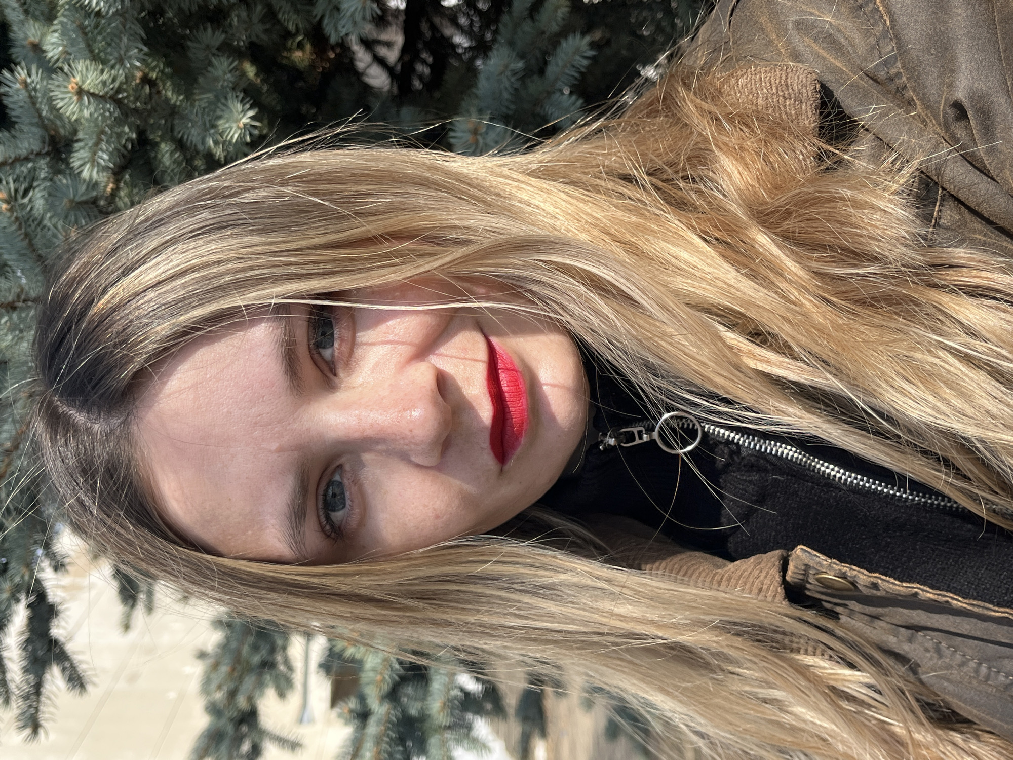 A portrait-style image, rotated 90 degrees counter-clockwise, features a young woman with long, flowing blonde hair that has darker roots. She has blue eyes, fair skin, and is wearing vibrant red lipstick. Her expression is neutral as she looks directly at the camera.
She is dressed in a dark, possibly black, inner garment, and an outer jacket in a shade of brown or tan. The jacket has a visible metallic zipper with a round pull tab, and a textured collar, possibly corduroy, which is visible near her shoulder.
The setting is outdoors on a bright, sunny day, evident from the strong highlights on her hair and face. The background consists of out-of-focus evergreen trees with green-blue needles, suggesting a park or natural area. A blurred, lighter expanse behind the trees might indicate snow-covered ground or a distant structure. The scene captures a direct portrait in what appears to be a cool climate, fitting the location context of Chaikovsky, Russia. No visible text is present in the image.