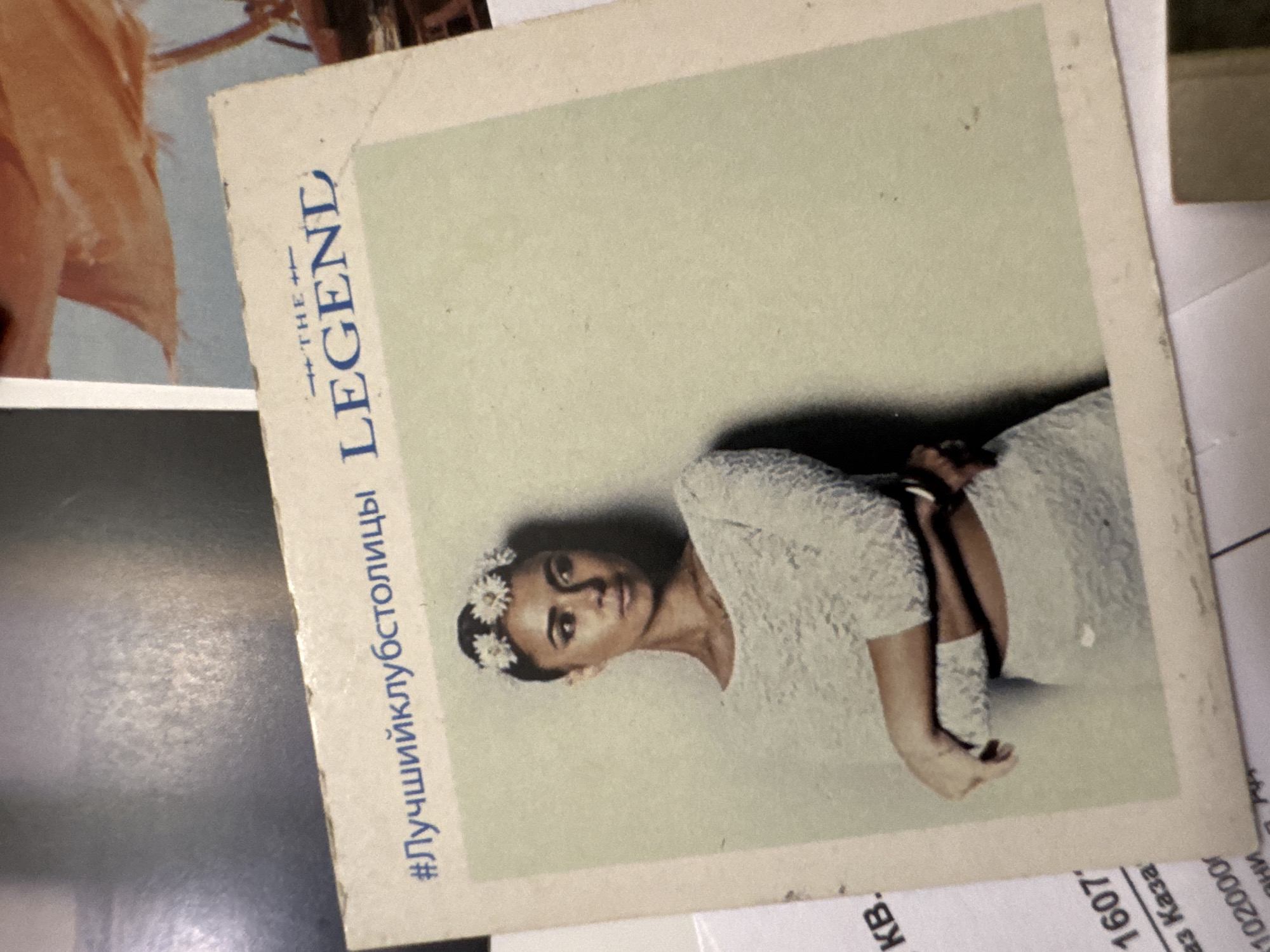 A color print of a woman is positioned against a backdrop of various papers and objects. The woman, with flowers in her hair, is wearing a light-colored garment. The printed text, "#Лучшийклубстолицы LEGEND" is visible on the side of the image. The woman's pose and attire suggest an advertisement or promotional material. The background objects include papers with Russian text. Without additional context, it's impossible to independently verify the location is Kazan, Russia, solely based on the provided information.