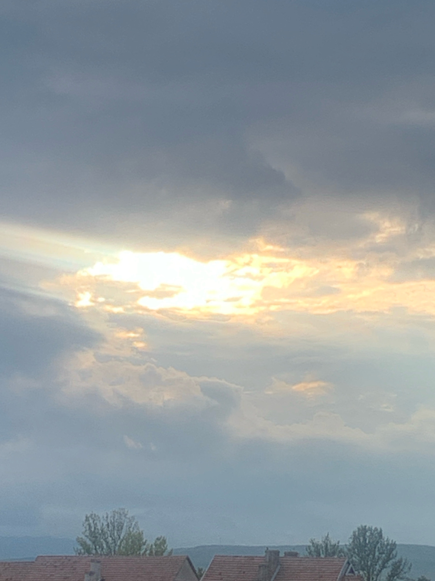 The image captures a cloudy sky with the sun partially visible, casting rays of light through the clouds. The lower portion of the image shows the tops of houses with tiled roofs and some trees, suggesting a residential area. The overall scene evokes a somewhat overcast or dusk-like atmosphere. The location is identified as Kolomyia, Ukraine.