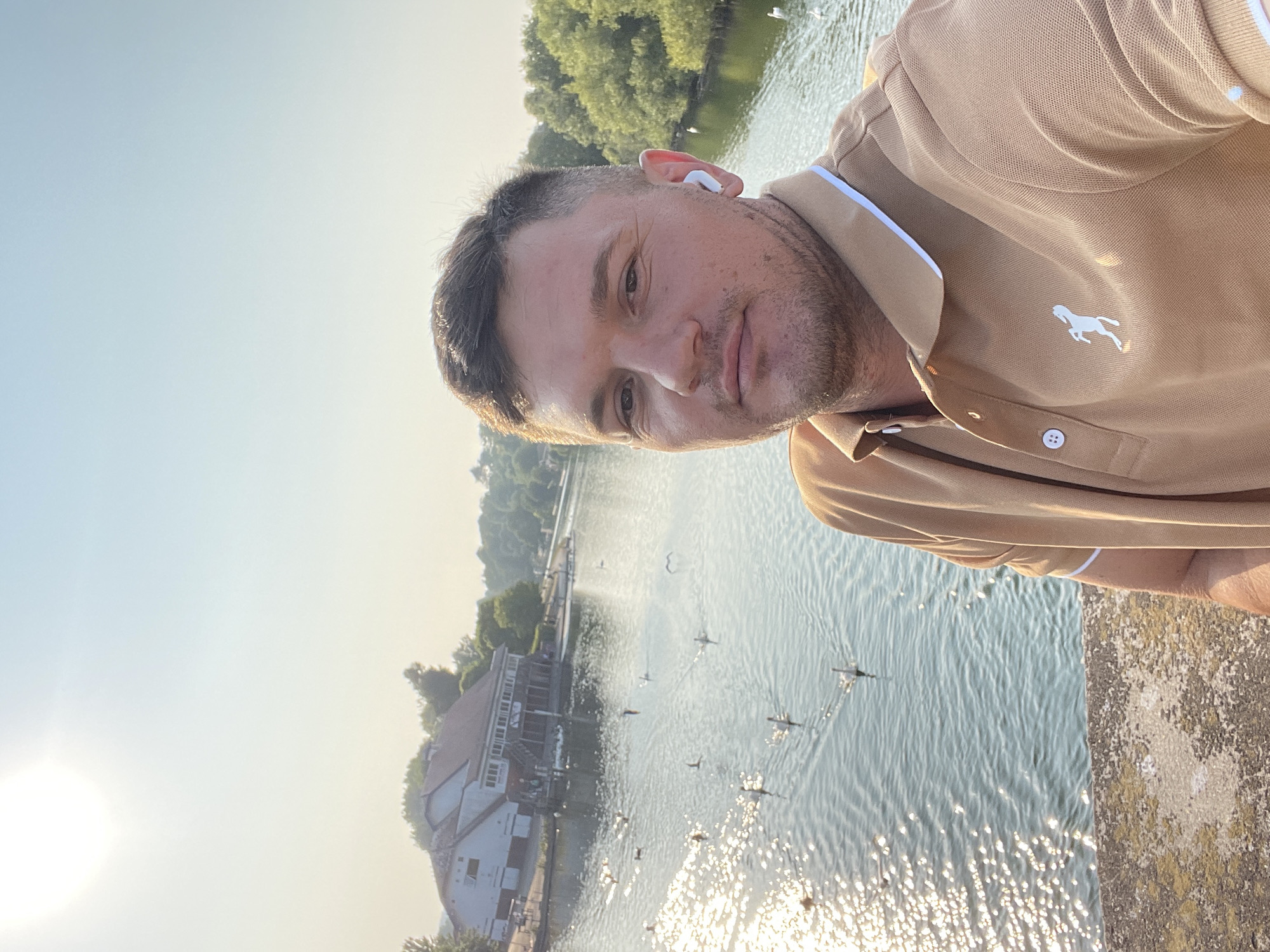 A man with short brown hair and a light tan polo shirt stands in the foreground, taking a selfie. He is wearing white wireless earbuds. In the background, a body of water reflects the sunlight. Several birds, possibly swans, are swimming on the water. Across the water, there is a building with a white facade and a dark roof, and lush green trees line the far bank of the water. The overall scene suggests an outdoor setting, possibly a park or a lake. The image appears to have been taken in the city of Reading, United Kingdom, given the presence of similar architecture and bodies of water.