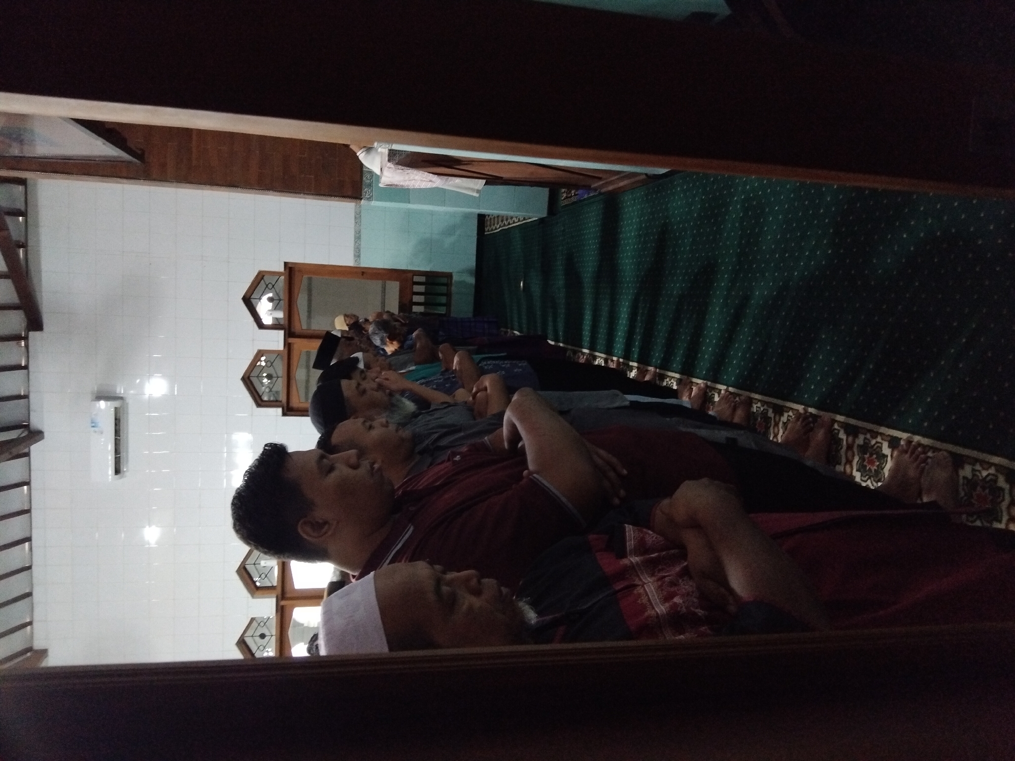 The image depicts a group of Muslim men standing in prayer at a mosque. The men are lined up in rows, facing a wooden structure that appears to be a minbar or a decorative wall. The mosque has a white tiled wall and a green carpeted floor with a traditional pattern. Natural light filters in from windows above the prayer area, and an air conditioning unit is visible on the wall. The overall atmosphere is one of solemnity and devotion. The presence of prayer mats and the attire of the men suggest a religious gathering. The architectural style and the humid, tropical environment hinted at by the lighting and materials could indicate a location in Southeast Asia, consistent with Karanganyar, Indonesia.