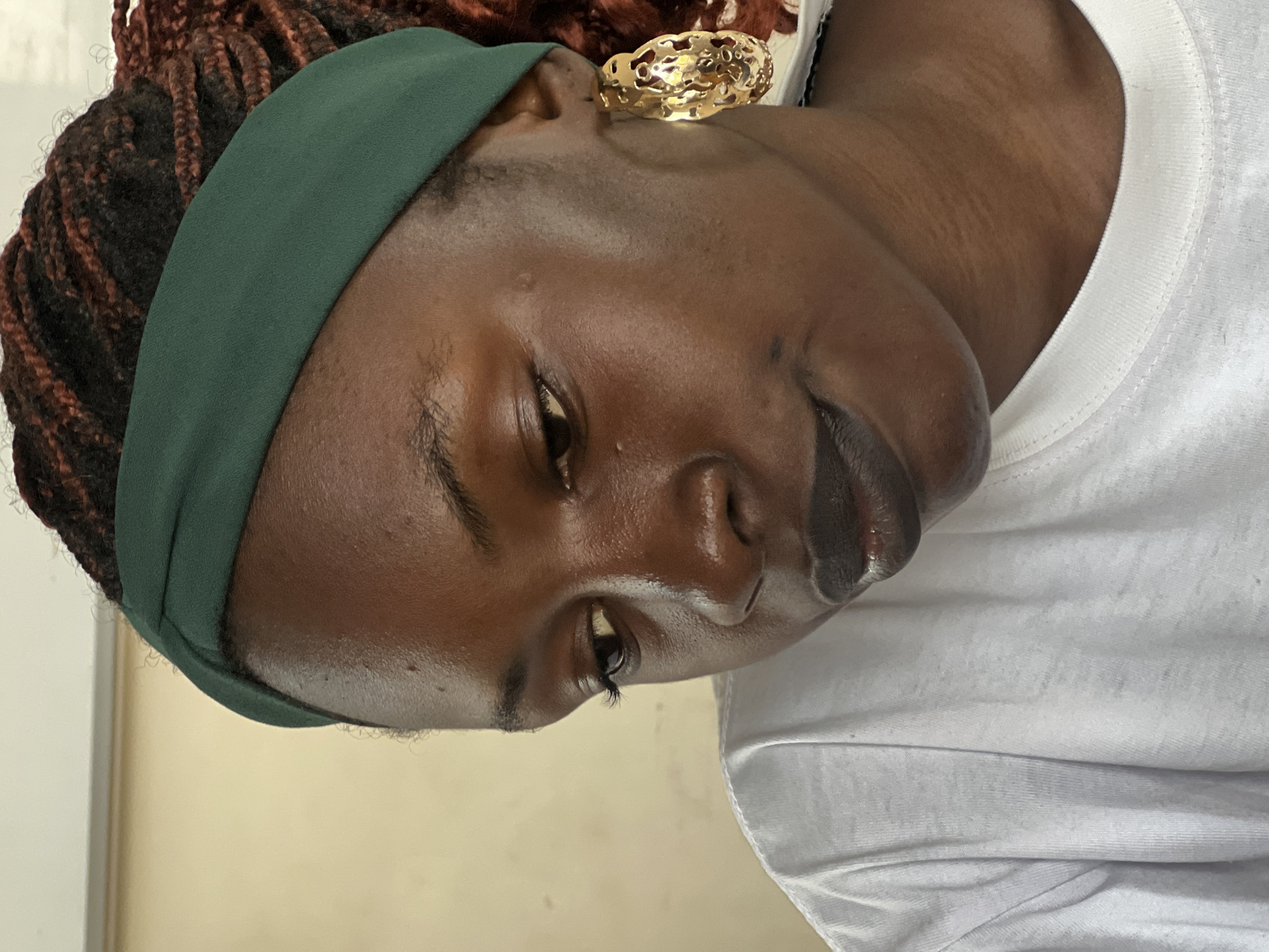 A close-up, head-and-shoulders, profile shot shows a young Black woman wearing a white t-shirt and a dark green headband. She has dark skin, braided hair with reddish highlights, and large, ornate gold hoop earrings. Her eyes are cast downward, and she has dark lipstick on. The background is a plain, light-colored wall, suggesting an indoor setting. No specific city details are visible.