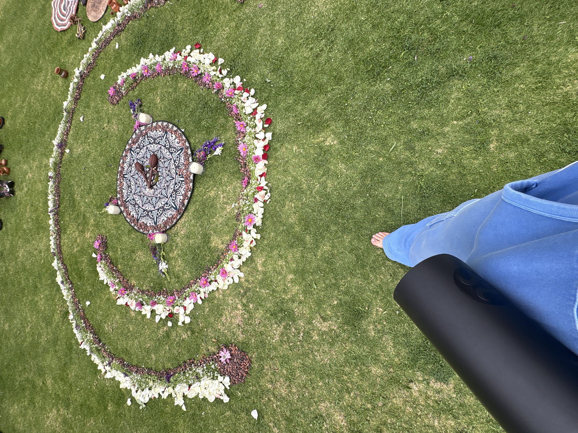 The scene is outdoors, on green grass. A person stands, barefoot, wearing blue clothing and holding a rolled-up black yoga mat. To the left, a spiral-shaped design is created using flowers and seeds, encircling a circular mandala-patterned mat with small objects placed on and around it. The scene appears to be in Santa Ana Acozautla, Mexico.