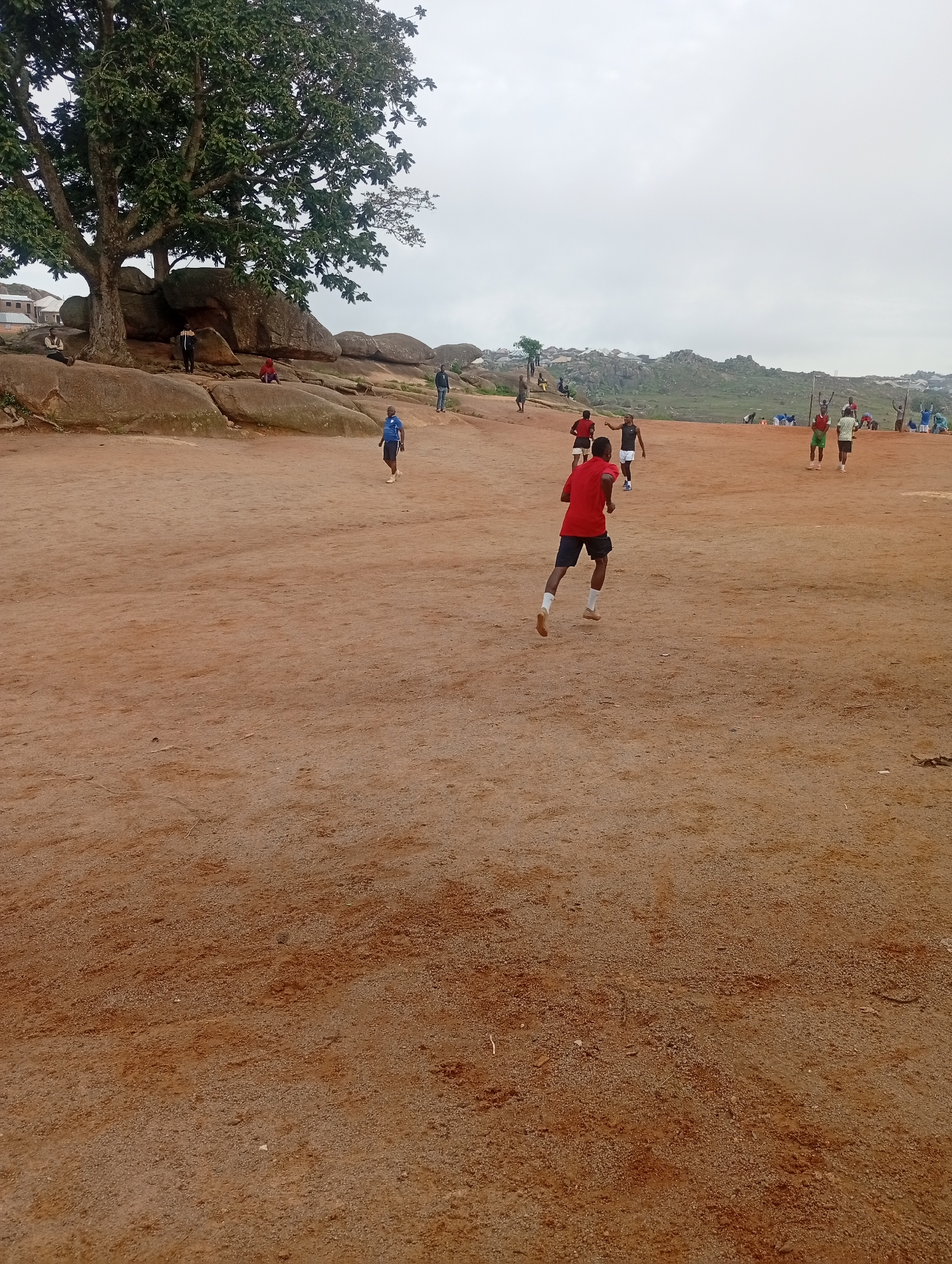 In an outdoor, arid environment, several individuals are engaged in physical activity on a reddish-brown dirt field. A large, mature tree with a sprawling canopy dominates the left side of the frame, its branches casting shadows over a cluster of large, weathered rocks. In the foreground, a person in a red shirt and dark shorts runs away from the camera. Further in the background, other individuals, some wearing athletic attire, are dispersed across the field. Some appear to be running or playing, while others stand and observe. The terrain is uneven and rocky, with hints of vegetation in the distance, suggesting a natural or undeveloped area within or on the outskirts of a settlement. The sky is overcast.