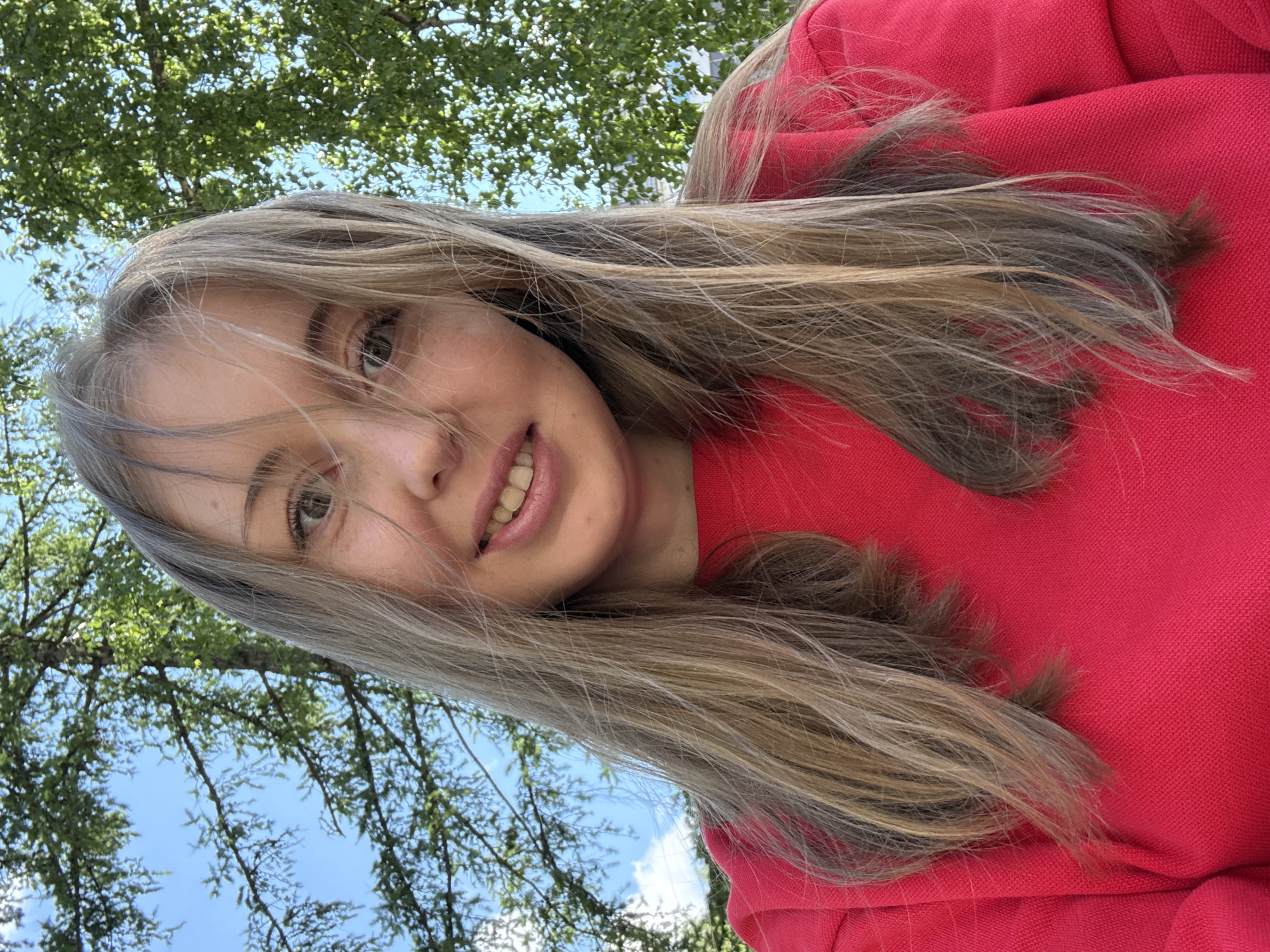 A young woman with long blonde-brown hair is centered in the image. She is wearing a red sweatshirt. The background consists of the branches and leaves of a tree against a blue sky. The location appears to be outdoors.