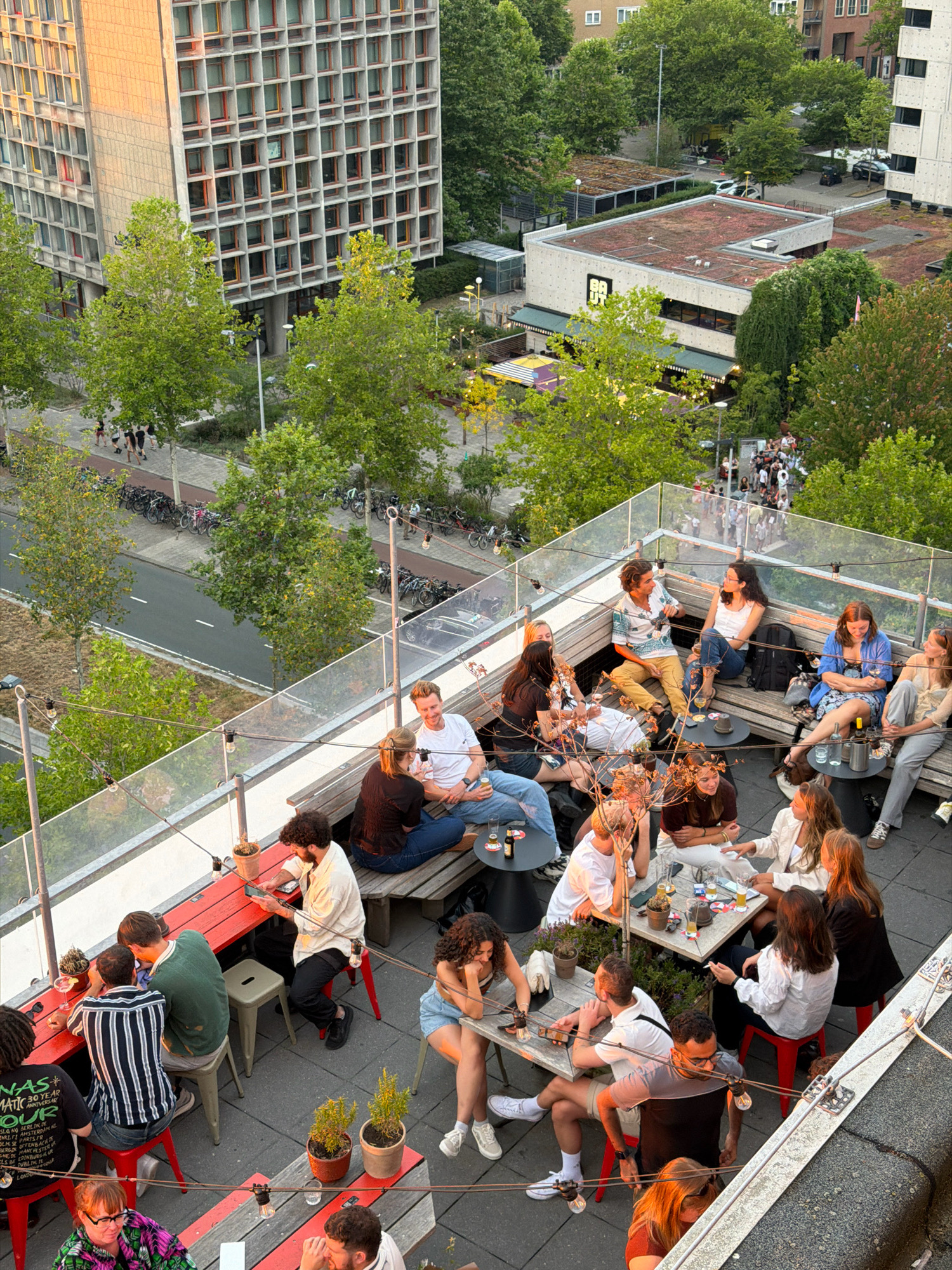 An aerial view captures a rooftop bar in Amsterdam, Netherlands, on a warm evening. The bar, situated on the top of a building, is filled with people socializing and enjoying drinks. The scene is bustling with activity, with groups of friends seated at tables, engaged in conversation and laughter. The atmosphere appears relaxed and convivial, with string lights casting a warm glow over the space. In the background, the cityscape of Amsterdam unfolds, with buildings and trees visible under the twilight sky. The image showcases a typical urban social gathering, highlighting the vibrant nightlife and community spirit of the city.
