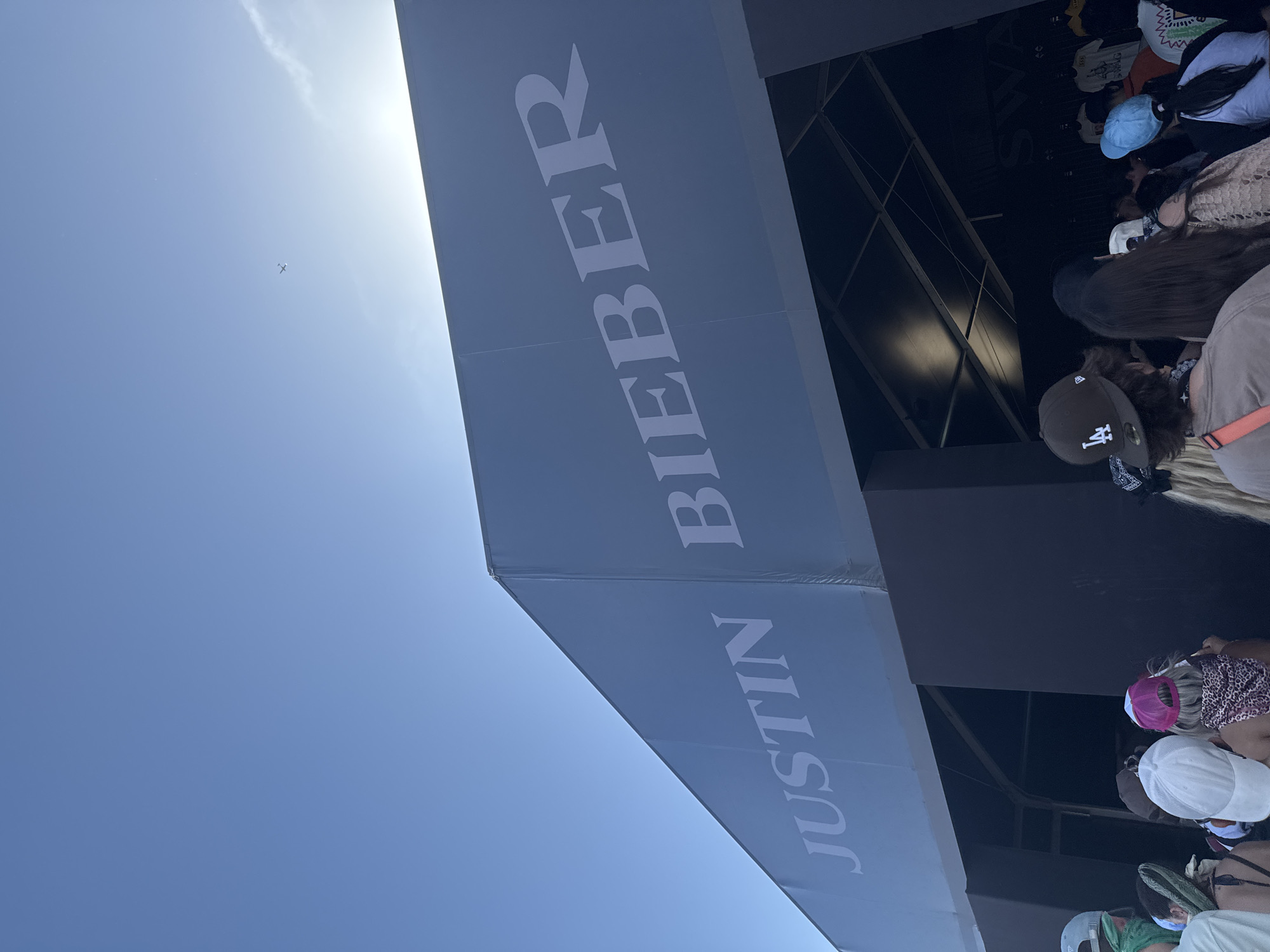 The image shows a banner that reads "JUSTIN BIEBER" against a bright blue sky with a visible sun. The banner is likely associated with a concert or event in Indio, United States of America, as suggested by the presence of a crowd of people, some wearing baseball caps, lining up beneath it. The architecture in the background indicates a commercial or event venue, and the light suggests daytime. The image conveys the atmosphere of a public event, potentially a concert, with the artist's name prominently displayed.