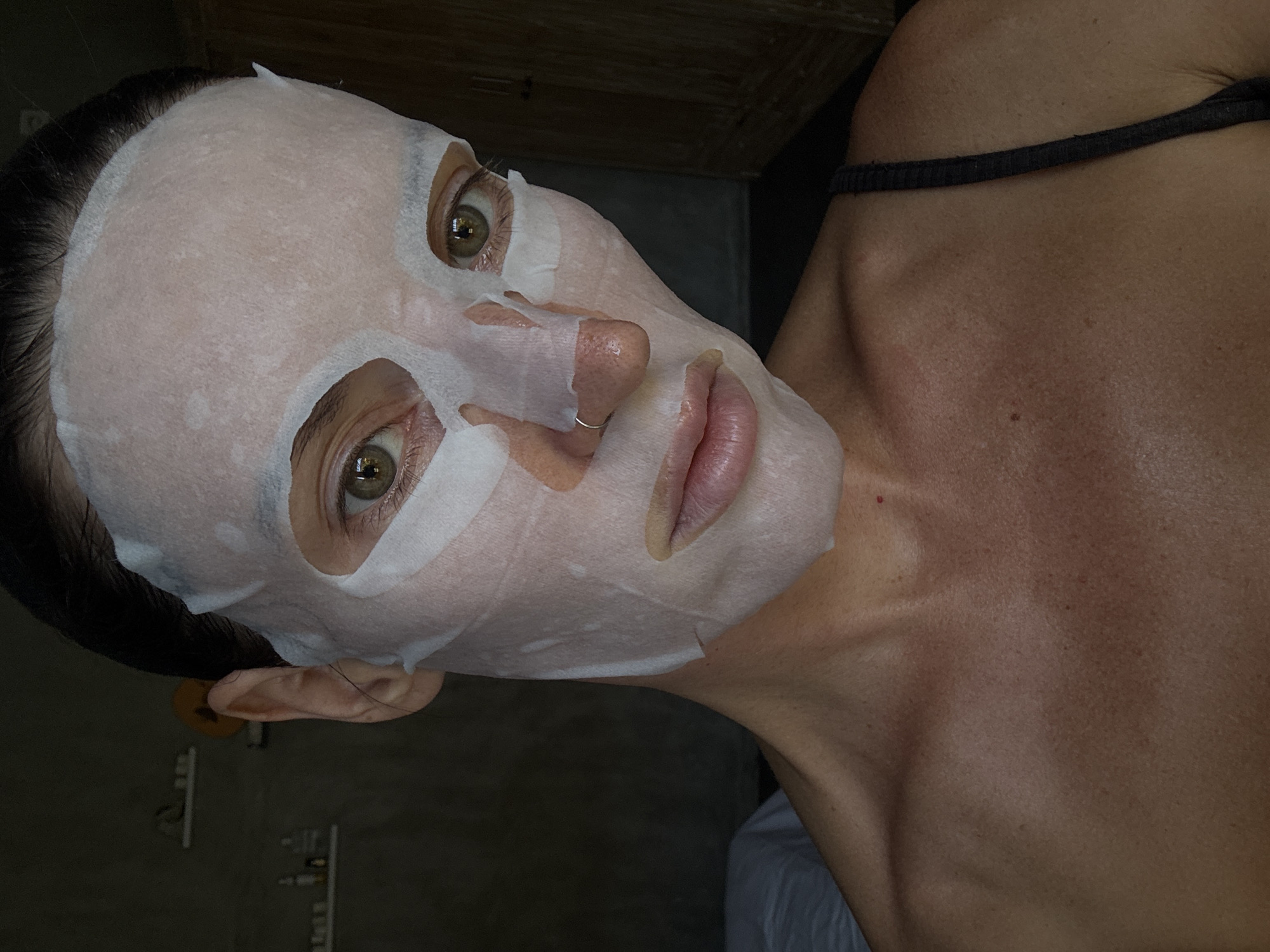 A woman is wearing a sheet mask on her face. The woman has light skin and light brown eyes. She has a nose ring on the right side of her nose. She has dark hair pulled back from her face. She is wearing a tank top. The photo was taken inside an indoor space with a dark background. The location may be in North Kuta, Indonesia.