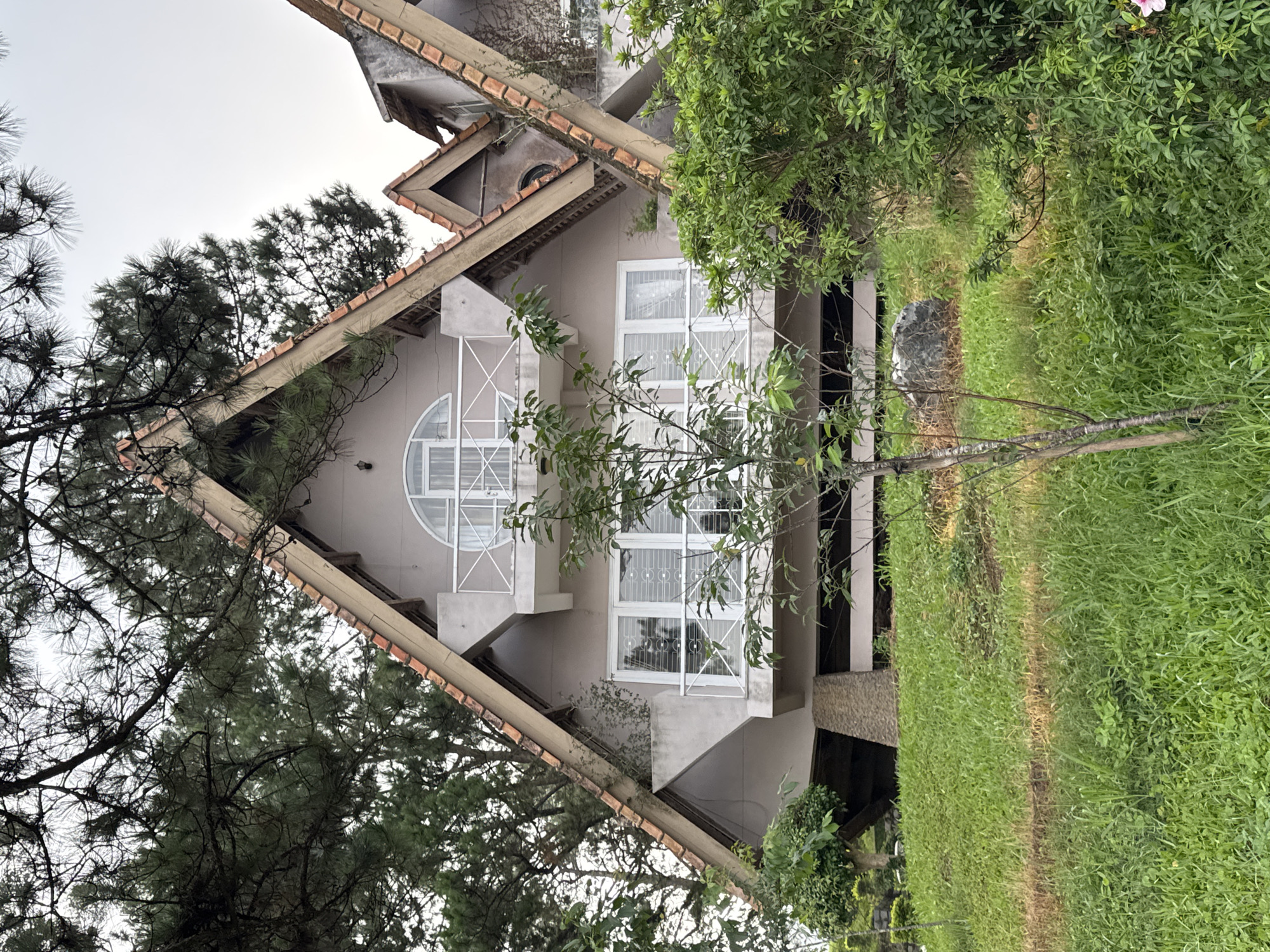 The image depicts a distinctive A-frame house situated on a grassy slope in Da Lat, Vietnam. The house features a light-colored facade with prominent triangular gables, adorned with red tile roofing. Large windows and balconies are visible, with a notable arched window on the upper level. The surrounding environment is lush and green, with dense trees and vegetation framing the structure. The sky is overcast. The scene appears to be a residential property, characterized by its unique architectural style and natural setting within the highlands of Da Lat. There are no people or animals visible in the image.