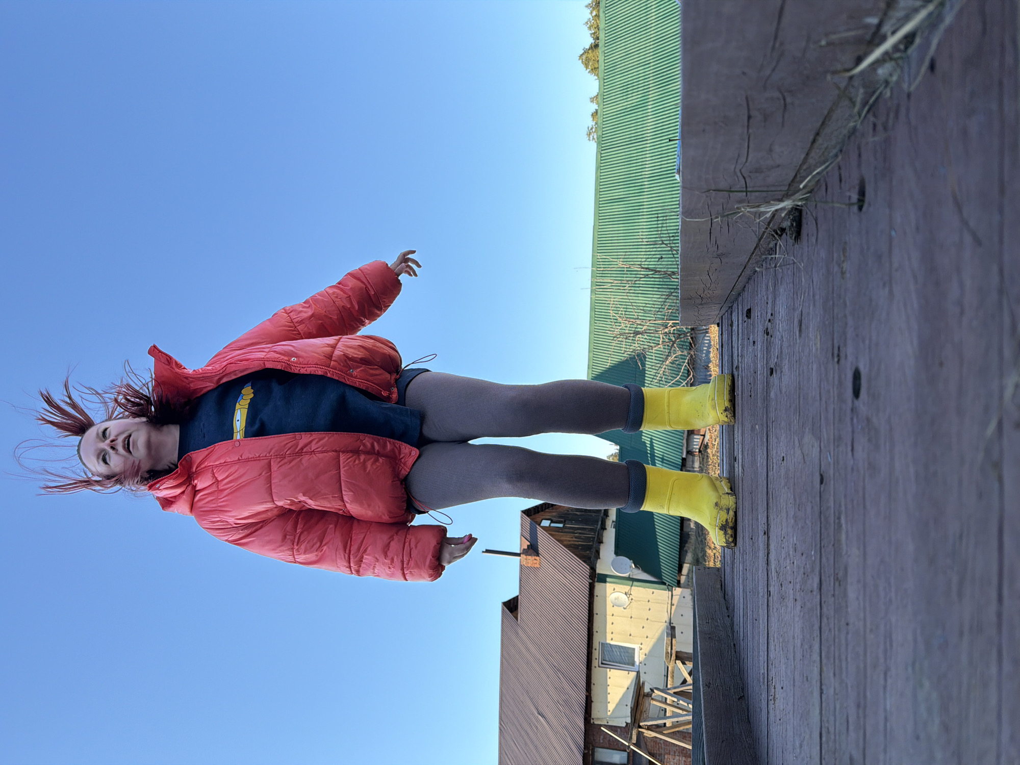 A young woman with red hair stands on a wooden platform, possibly a deck or boardwalk. She is wearing a coral-colored puffer jacket, a navy blue shirt with a yellow graphic, gray leggings, and yellow rain boots. She has one arm outstretched toward the bright blue sky, and her hair is blowing in the wind. The platform appears to be elevated, with a green corrugated metal roof visible in the background. Behind the platform, there's a partially obscured view of a building or buildings, some with brown roofs. The scene is outdoors, with clear, sunny weather. The specific location in Chaikovsky, Russia, cannot be determined from the visual data provided.