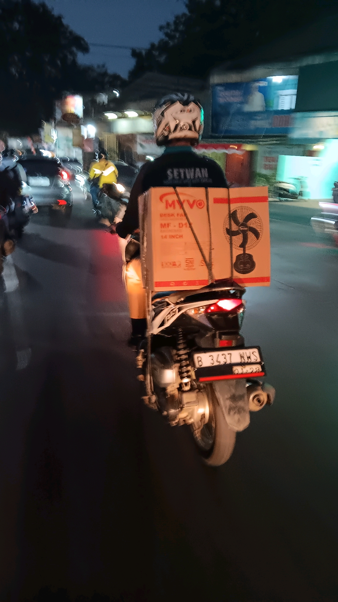 The image shows a delivery person on a scooter at night on a street in South Tangerang, Indonesia. The delivery person is wearing a helmet and a jacket with "SETWAN" written on the back. They are carrying a large cardboard box containing a fan. The box has "MYVO" printed on it. The scooter has a license plate that reads "B 3437 NWS". In the background, other vehicles and streetlights are visible. The scene appears to be a busy street with moderate traffic.