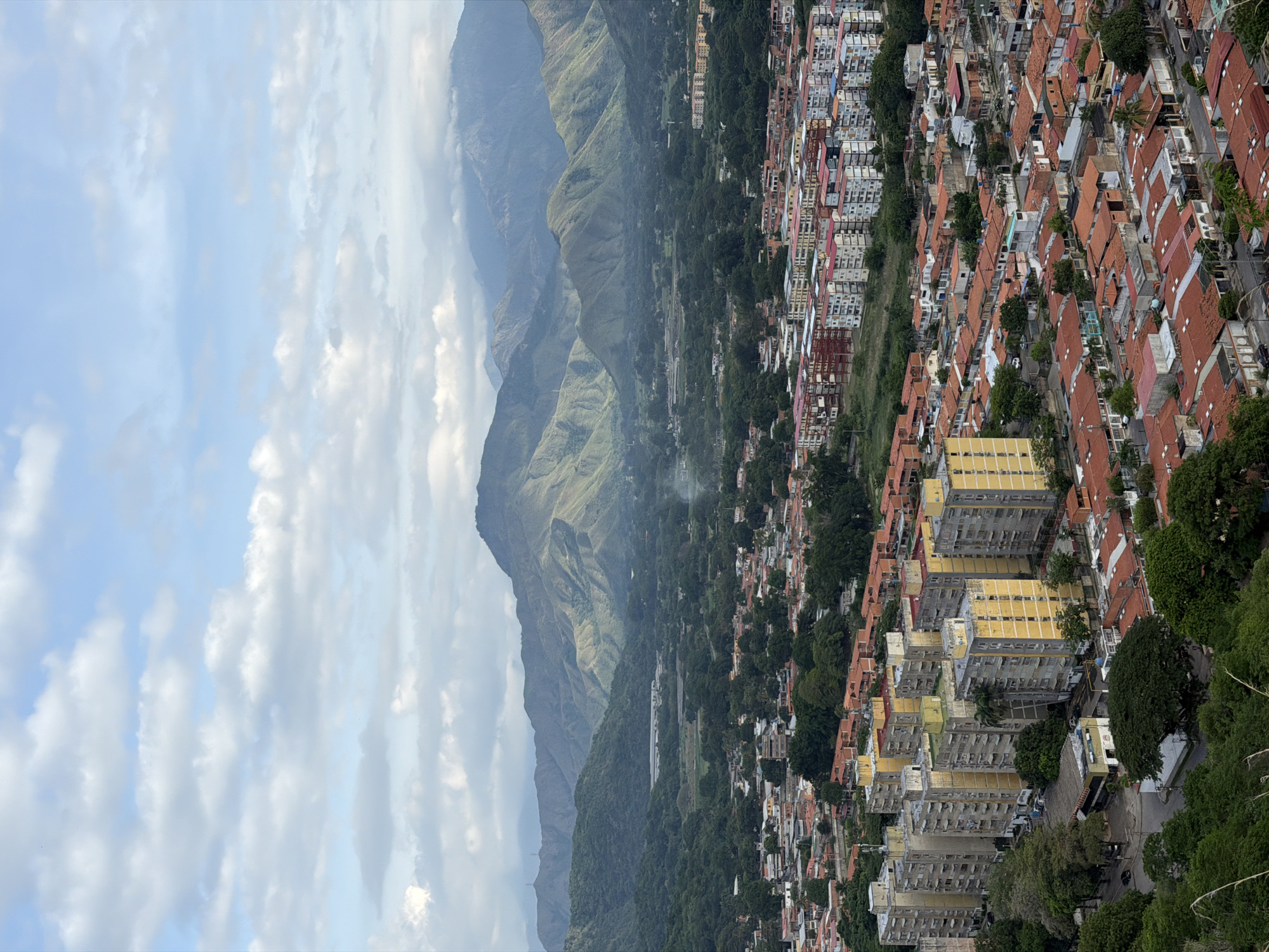This is a wide-angle, elevated landscape photograph of a densely populated urban area at the base of a mountain range, likely in Turmero, Venezuela. The sky above is a mix of light blue with scattered white clouds. In the foreground, a cluster of residential buildings, predominantly low-rise houses with red tile roofs, dominate the view. These are interspersed with taller apartment buildings, some with yellow facades, others with a mix of white and colorful exteriors. Trees and vegetation are visible throughout the residential areas, with a more dense forested area in the lower left corner.
The middle ground shows a transition from residential areas to a flatter expanse with more dispersed buildings, some appearing to be industrial or commercial structures, possibly including greenhouses. Further back, the landscape rises into rolling hills covered in lush green vegetation, which gradually merge into the imposing, layered mountain range that forms the background. The mountains are varied in height and gradient, with some peaks appearing hazy in the distance. The overall impression is of a city nestled within a natural, mountainous environment. There are no visible people or distinct actions taking place. The scene appears to be captured during daylight, likely late afternoon or early morning, given the soft lighting.