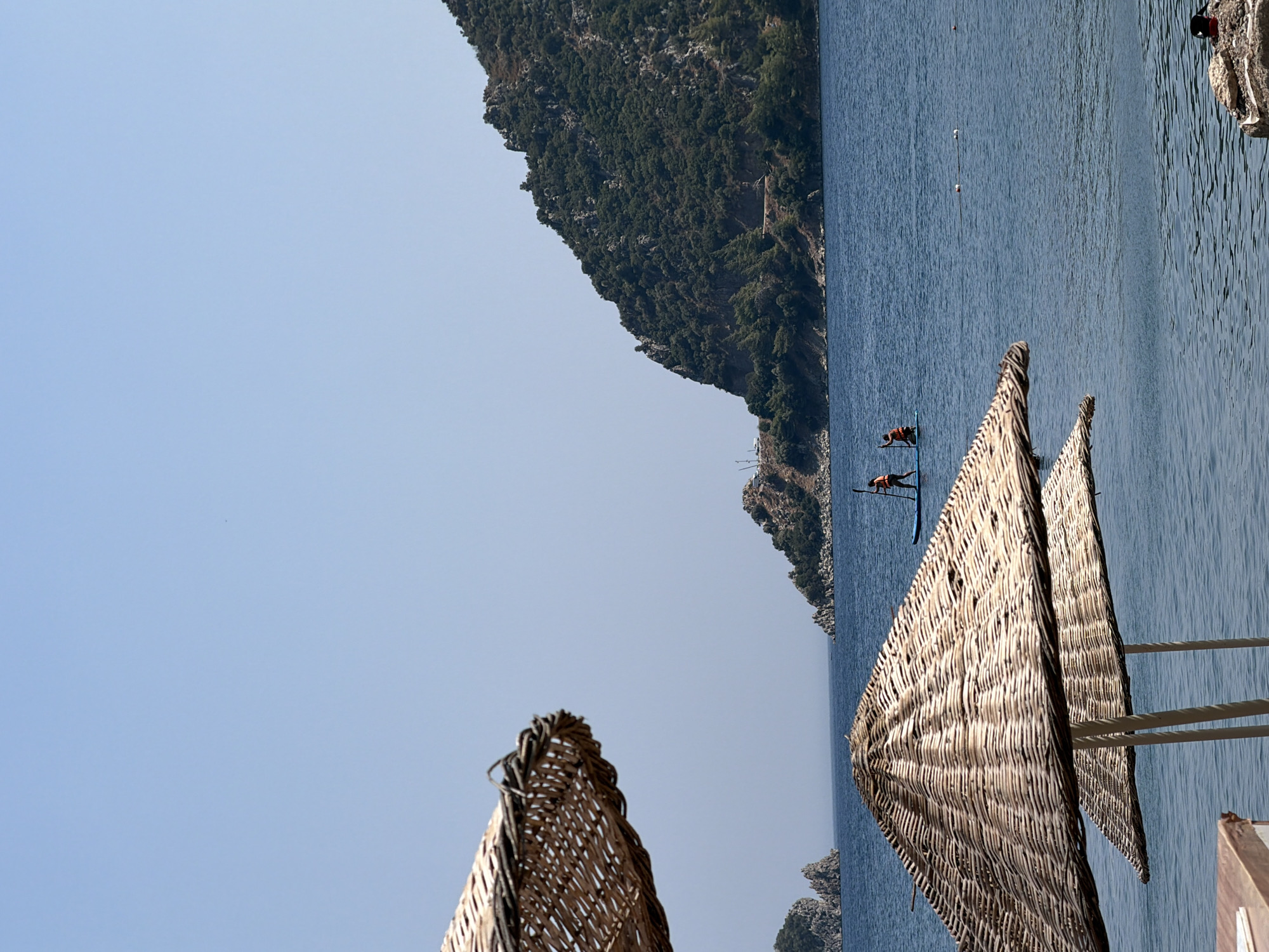 Two people are paddleboarding on a calm, blue sea under a clear sky. They are positioned in the middle ground of the image, with a wooded hillside rising in the background and the sea stretching out before it. In the foreground, two straw beach umbrellas are visible, partially obscuring the view of the water. One umbrella is larger and closer to the viewer, while the second is smaller and further back. The water is calm, with gentle ripples. The overall scene depicts a peaceful day at the beach in Marmaris, Turkey.