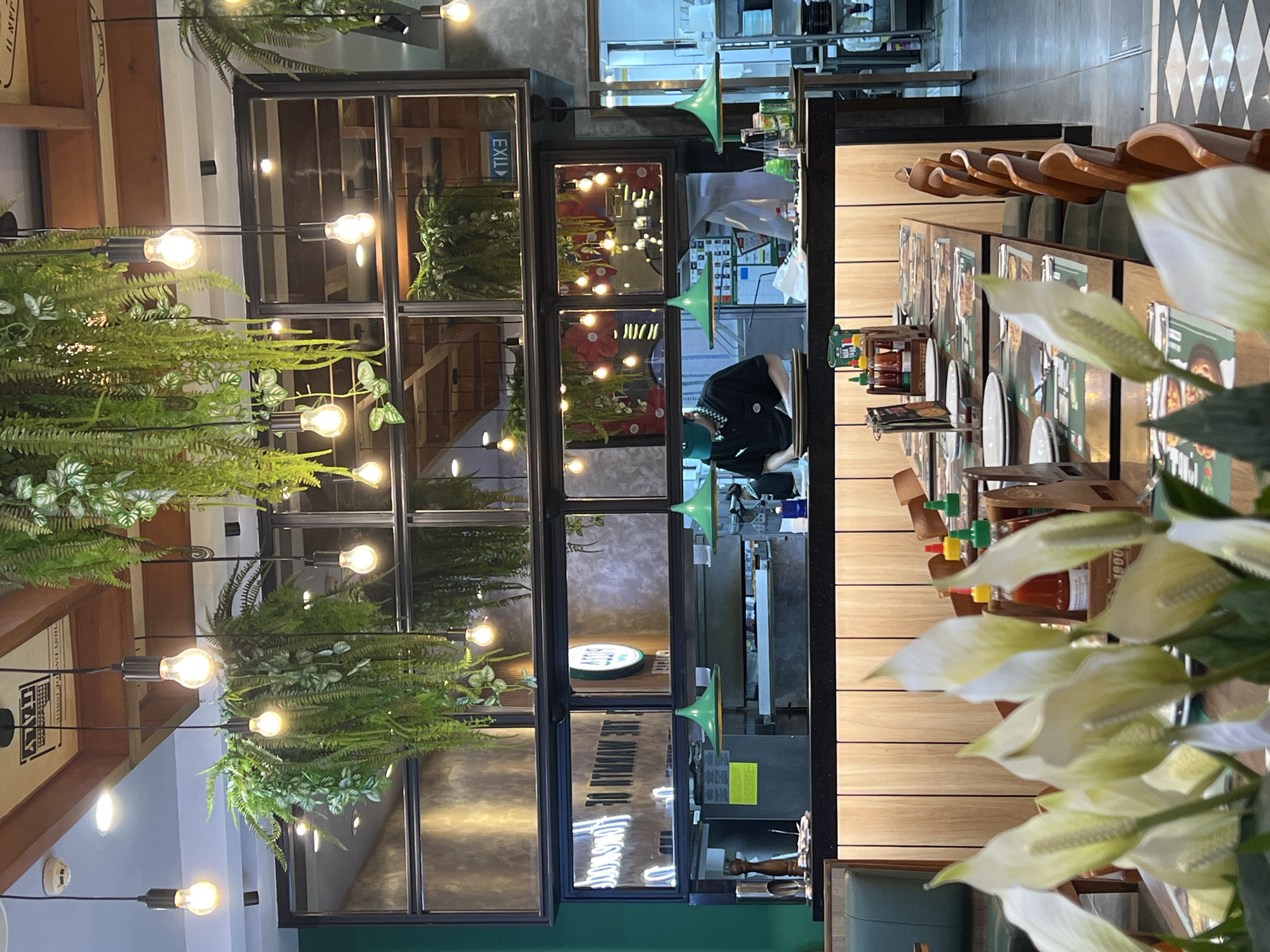 The image depicts the interior of a restaurant, possibly in Đà Nẵng, Vietnam. In the foreground are white flowers. Behind those are several tables set with plates, silverware, menus, and condiments such as hot sauce. The chairs are made of light-colored wood. In the background is a mirrored partition with plants and decorative lightbulbs. Behind that appears to be a food preparation area with a worker wearing a mask. The floor in the back is tiled.