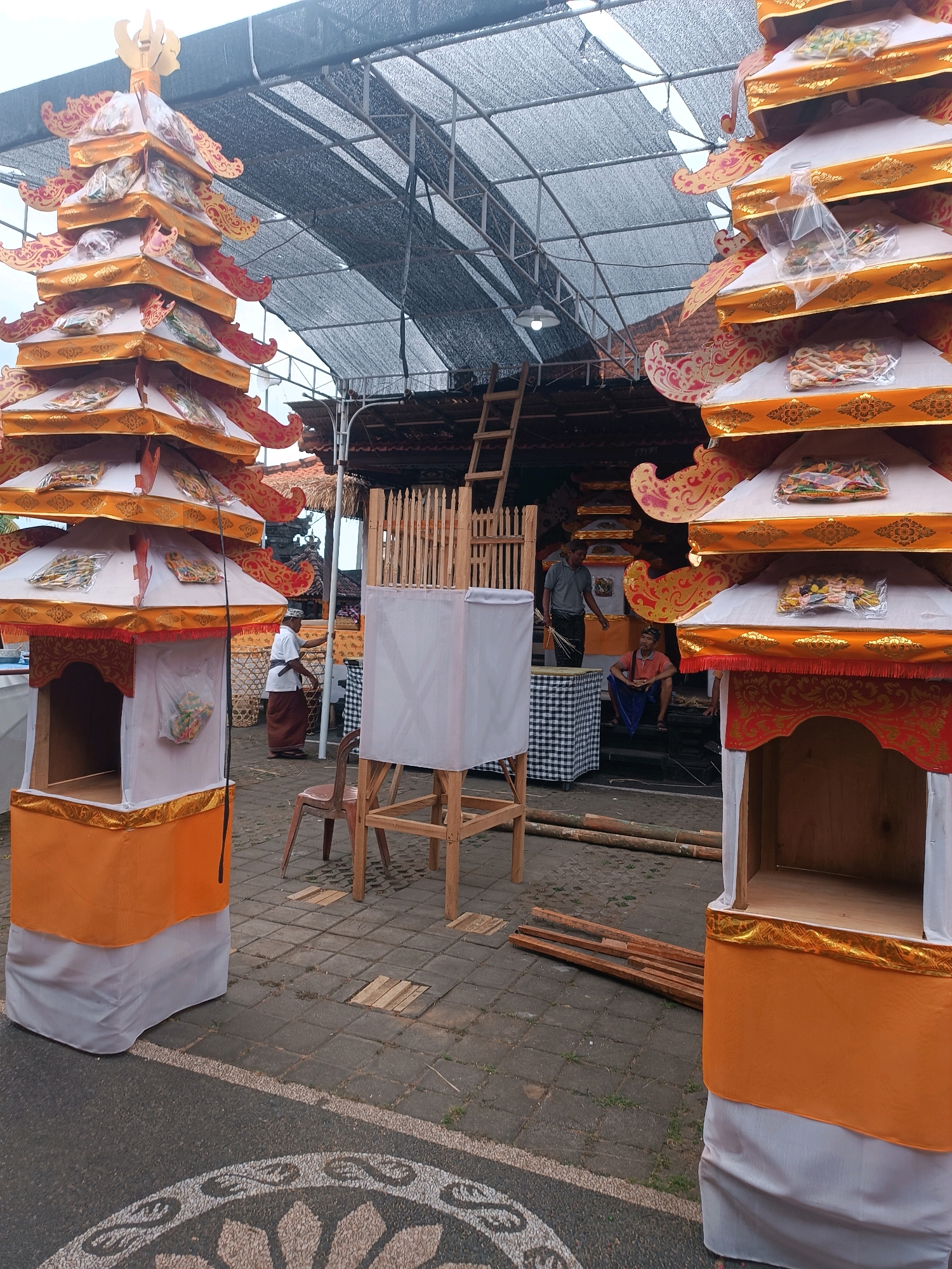 The image depicts an outdoor scene in Selemadeg, Indonesia, likely a public space or a temple complex, under a large, dark mesh canopy that provides shade. In the foreground, two elaborately decorated structures, resembling tiered towers or shrines, are prominently displayed. These structures are adorned with orange and white fabric, gold trim, and ornamental red and gold decorative motifs. They appear to be made of wood with fabric coverings. The tiered tops of these structures contain items wrapped in clear plastic.
To the left, a man in traditional Balinese attire, a white shirt and a brown sarong, is standing near one of the structures, appearing to be engaged in some preparation or activity. To the right, another identical structure stands.
In the middle ground, a white, cubical object covered with a white fabric sits on a wooden frame. Further back, there are wooden planks and scaffolding, suggesting ongoing construction or setup. Several people are visible in the background: one man in a grey shirt and dark pants is standing, holding what appear to be dried palm leaves or similar materials, near a checkered black and white cloth. Another person, wearing an orange shirt and blue pants, is seated. The ground is paved with rectangular stone tiles, with a decorative circular mosaic pattern visible on the asphalt road in the lower part of the image. The overall impression is of a festive or ceremonial preparation, possibly for a local event or religious ceremony.