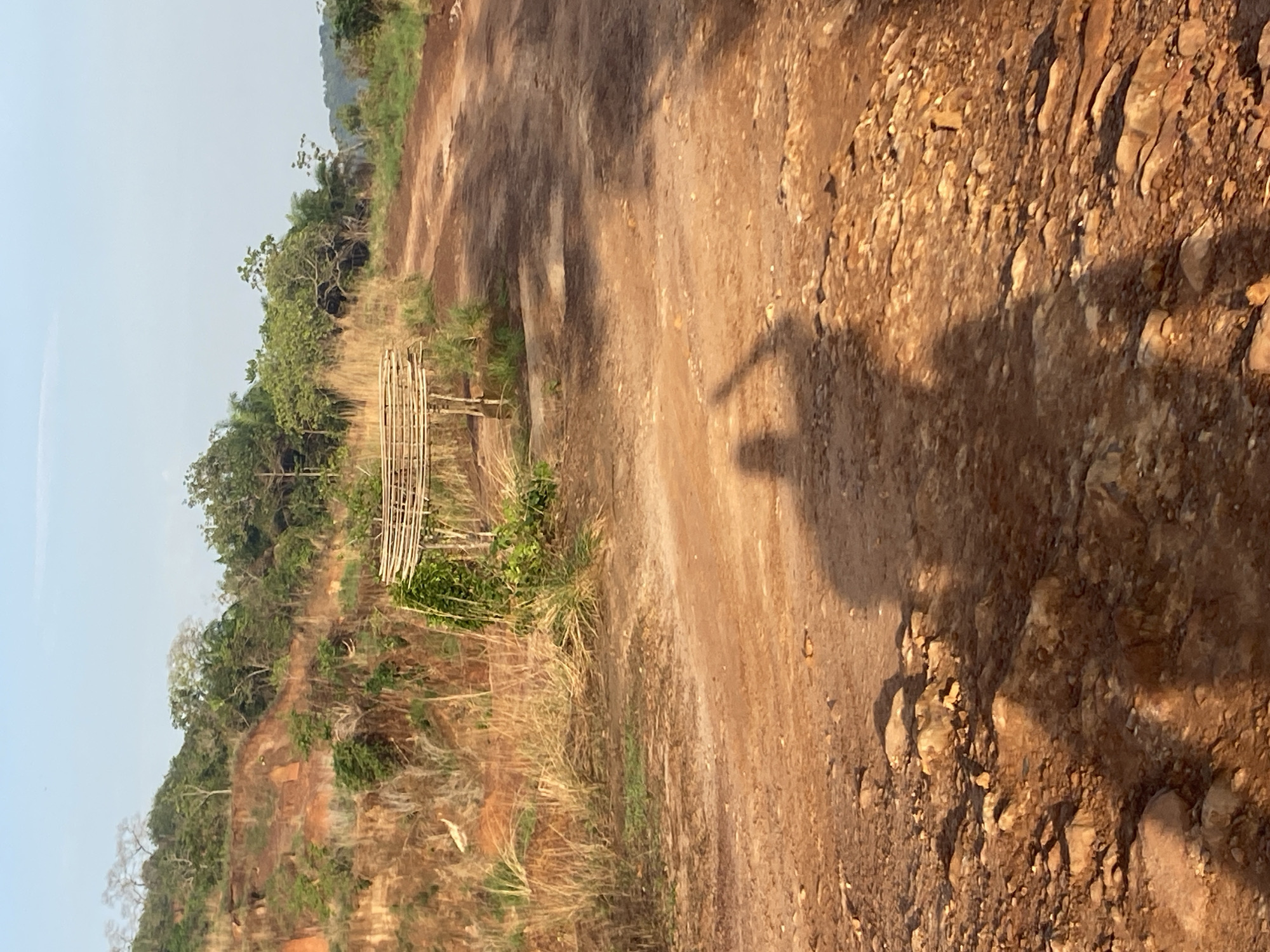 The image depicts a rural scene in Amancho, Nigeria. The foreground features a dirt road covered with loose rocks and soil. A shadow of a person is visible on the road, their arm raised in a possible greeting. To the left of the road, the land rises, covered in dried grasses, sparse vegetation, and trees. A makeshift wooden structure, possibly a bench or small platform, is situated among the vegetation. The sky is pale and clear.