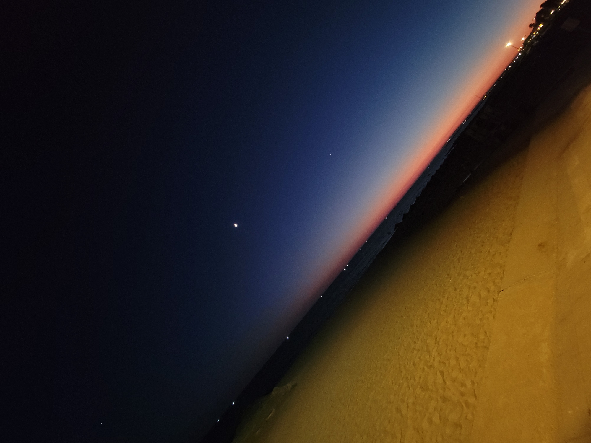 A tilted, wide-angle, long-exposure photograph captures a beach scene in Quarteira, Portugal, during twilight. The foreground is dominated by a sandy beach bathed in warm, artificial light, which casts long shadows and highlights the texture of the sand. The middle ground features the dark, calm sea stretching towards the horizon. The sky transitions from a deep blue at the top to a soft gradient of pink and orange near the horizon, indicative of the sun having recently set. A crescent moon is visible in the upper portion of the sky, along with several faint stars and scattered lights along the distant coastline, suggesting the presence of buildings and streetlights in the city. The image is devoid of any people or animals, presenting a serene and empty coastal landscape.