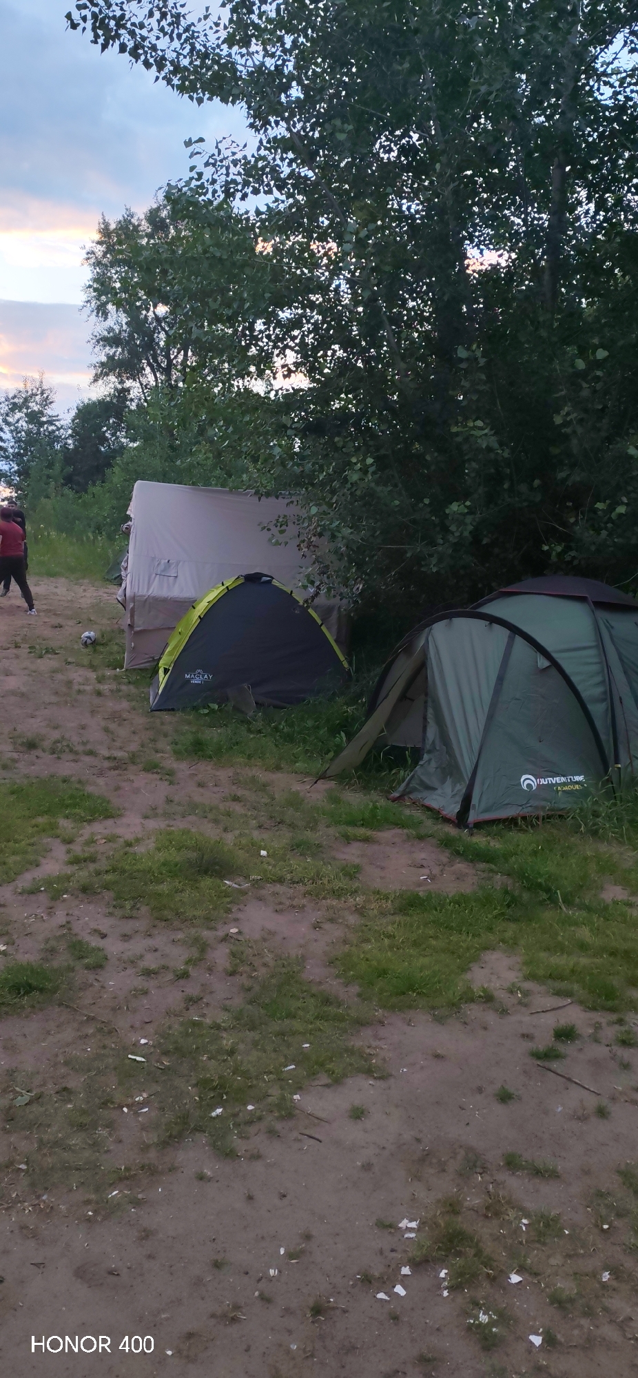 The image depicts an outdoor campsite scene in Noviy, Russia, featuring multiple tents set up on a dirt and grass area. In the foreground, a green and black dome tent is visible with the brand name "MACLAY" printed on it. To the right, another larger green tent, partially obscured by trees, has the brand name "OUTVENTURE" visible. In the background, a larger, light-colored tent or structure is set up, and two individuals in red and dark clothing are standing near it. The environment includes lush green trees and foliage surrounding the campsite, with a cloudy sky overhead, suggesting it might be late afternoon or evening. The ground is a mix of soil and patchy grass, with some debris scattered around.