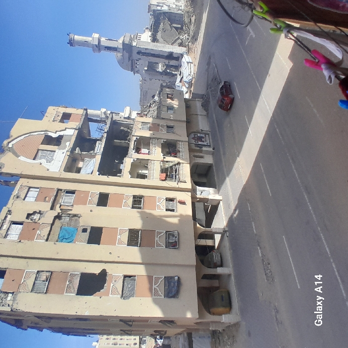 The image displays a severely damaged urban street scene in Khan Yunis, Palestinian Territory, under a clear blue sky. A prominent multi-story building, appearing to be residential or mixed-use, occupies the left and central mid-ground. Its facade shows extensive structural damage, including large sections of walls blown out, numerous shattered or missing windows, and visible impact marks consistent with shelling or gunfire. Some window openings are covered with blue or black tarpaulins and blankets. The ground floor features an open arcade structure, under which at least two parked cars, one yellow and one silver/grey, are partially visible.
To the right and rear of this building, a large mosque with a distinct minaret and dome is also significantly damaged, with substantial portions of its structure collapsed and surrounded by rubble. An asphalt road with white lane markings runs horizontally across the scene. A red hatchback car is parked or idling on the right lane of this road, facing away from the viewer. Further damaged buildings and debris are visible in the distant background. The photograph is taken from an elevated perspective, with a clothesline and several clothes pegs partially visible in the bottom right foreground. No individuals are discernible in the scene.